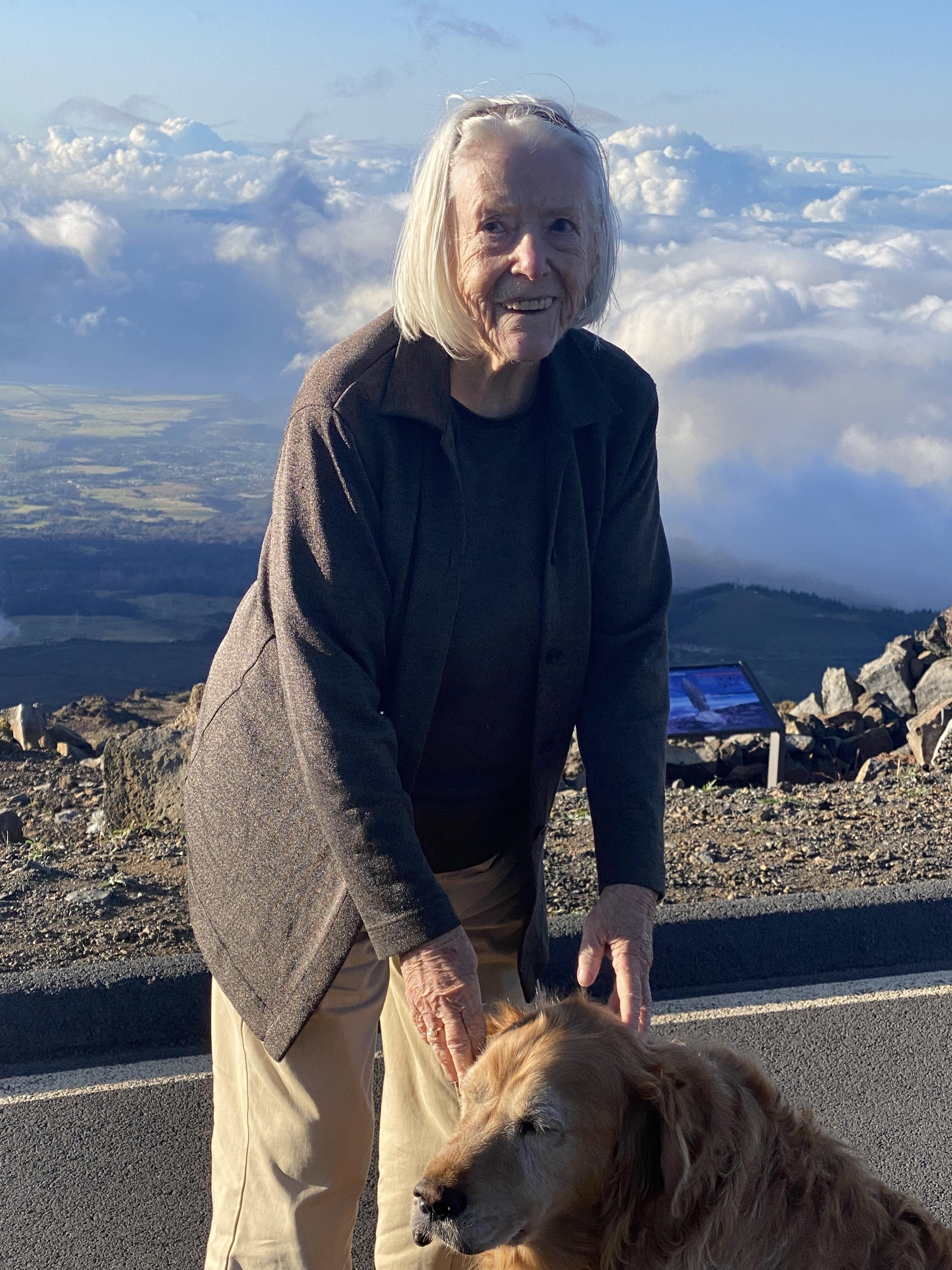 An elderly woman enjoys a moment with her golden retriever on a mountain surrounded by clouds.