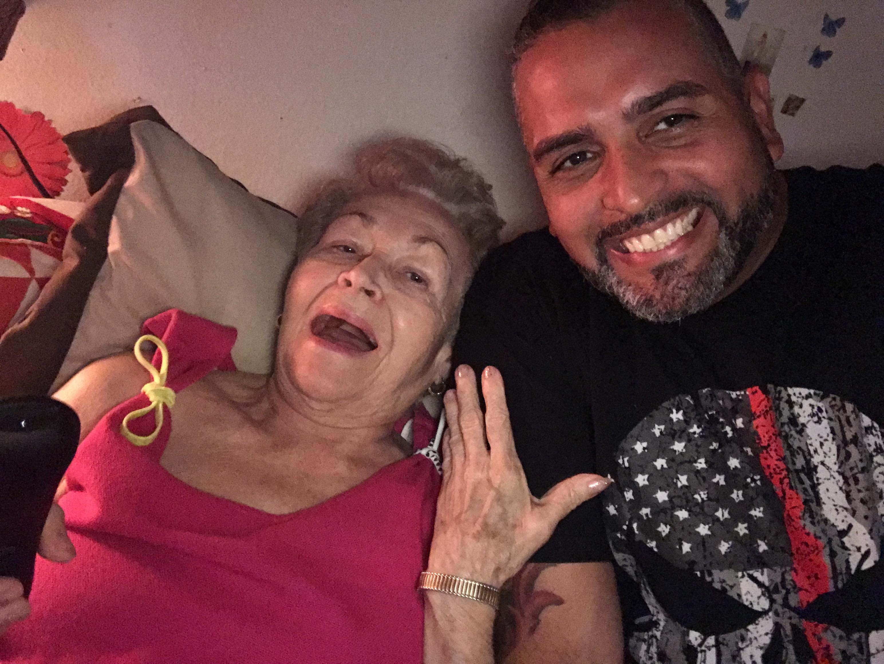 A man smiles joyfully next to an elderly woman, both enjoying a warm moment together indoors.