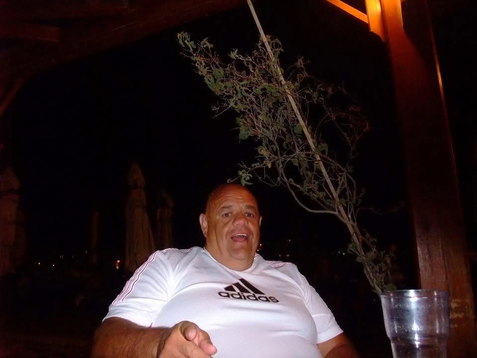 A man enjoys a drink under the warm glow of outdoor lighting, surrounded by trees.