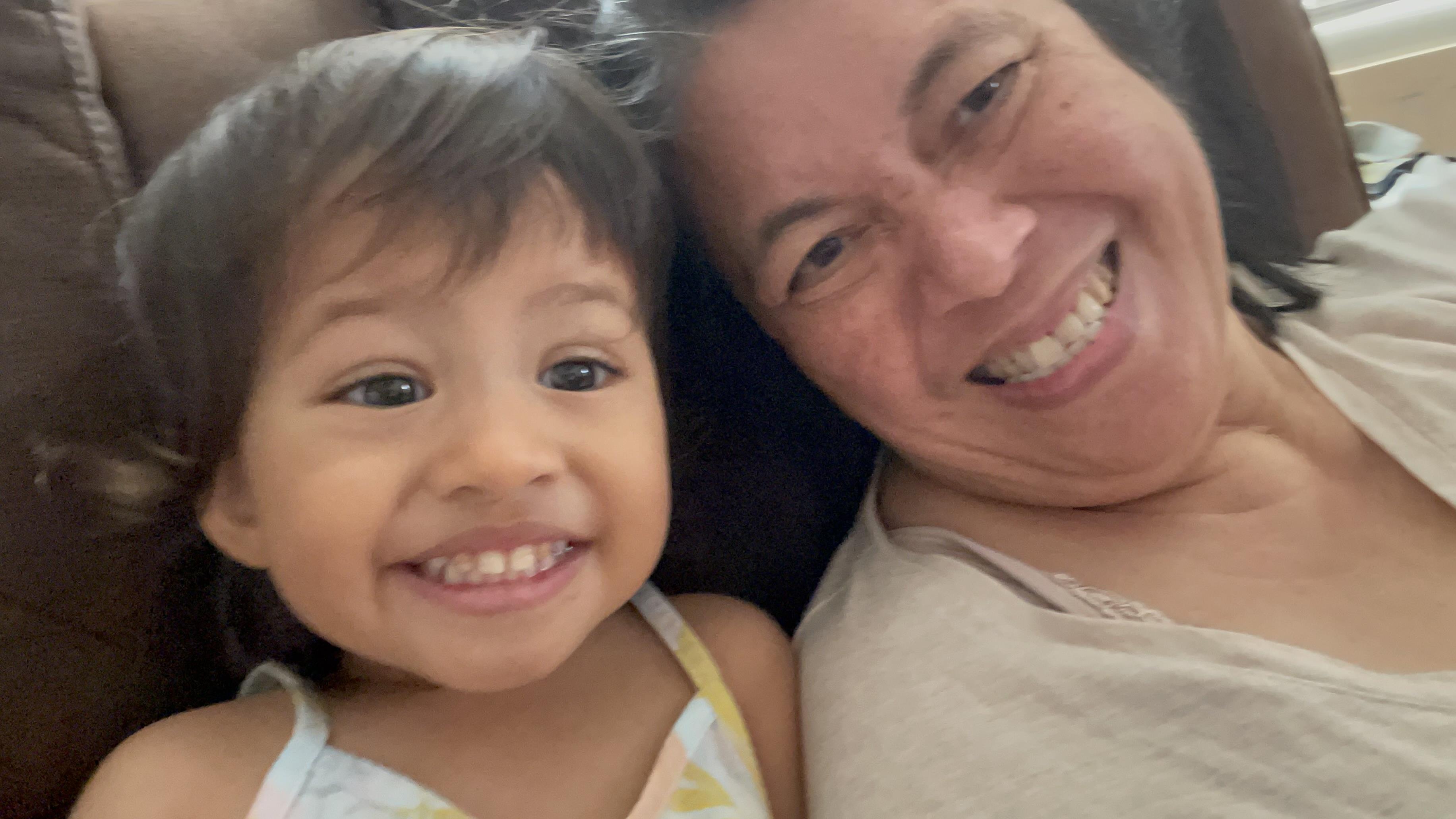 A grandmother smiles broadly alongside her cheerful granddaughter while sitting on a cozy couch.