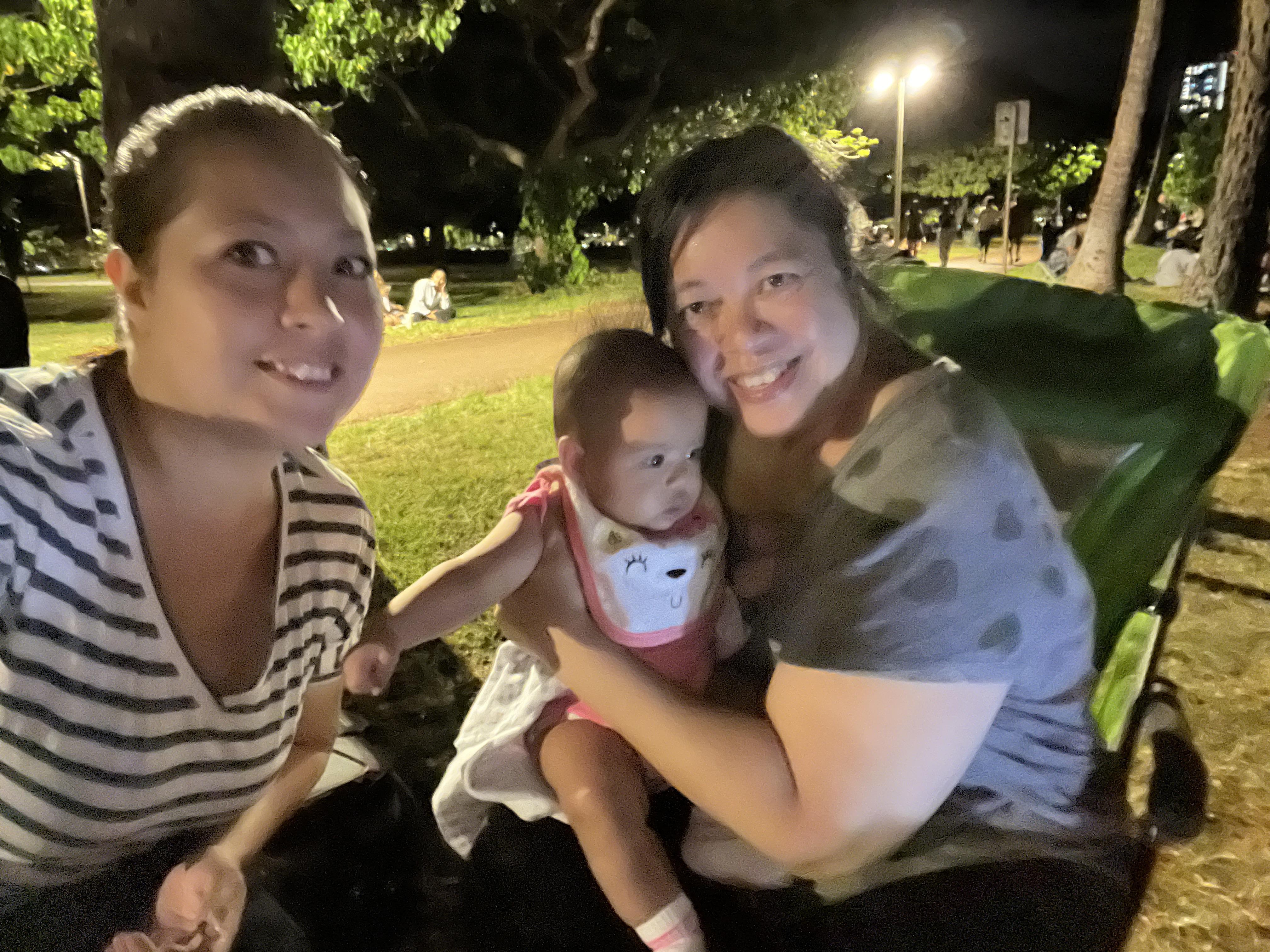 Two friends smile while holding a baby, surrounded by a lively park atmosphere at night.
