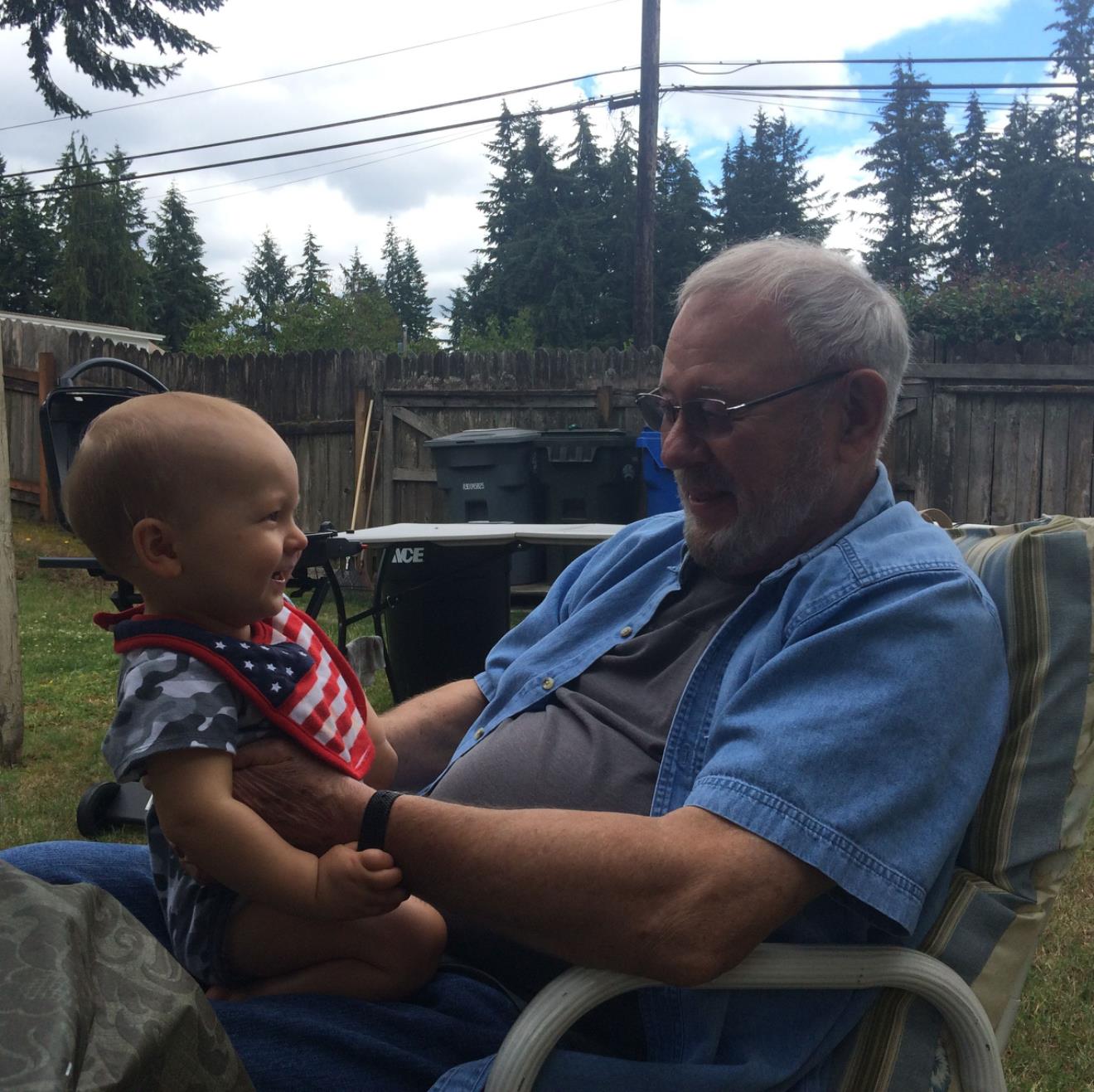 Elderly man smiles at laughing baby during a warm day outside in a backyard setting.