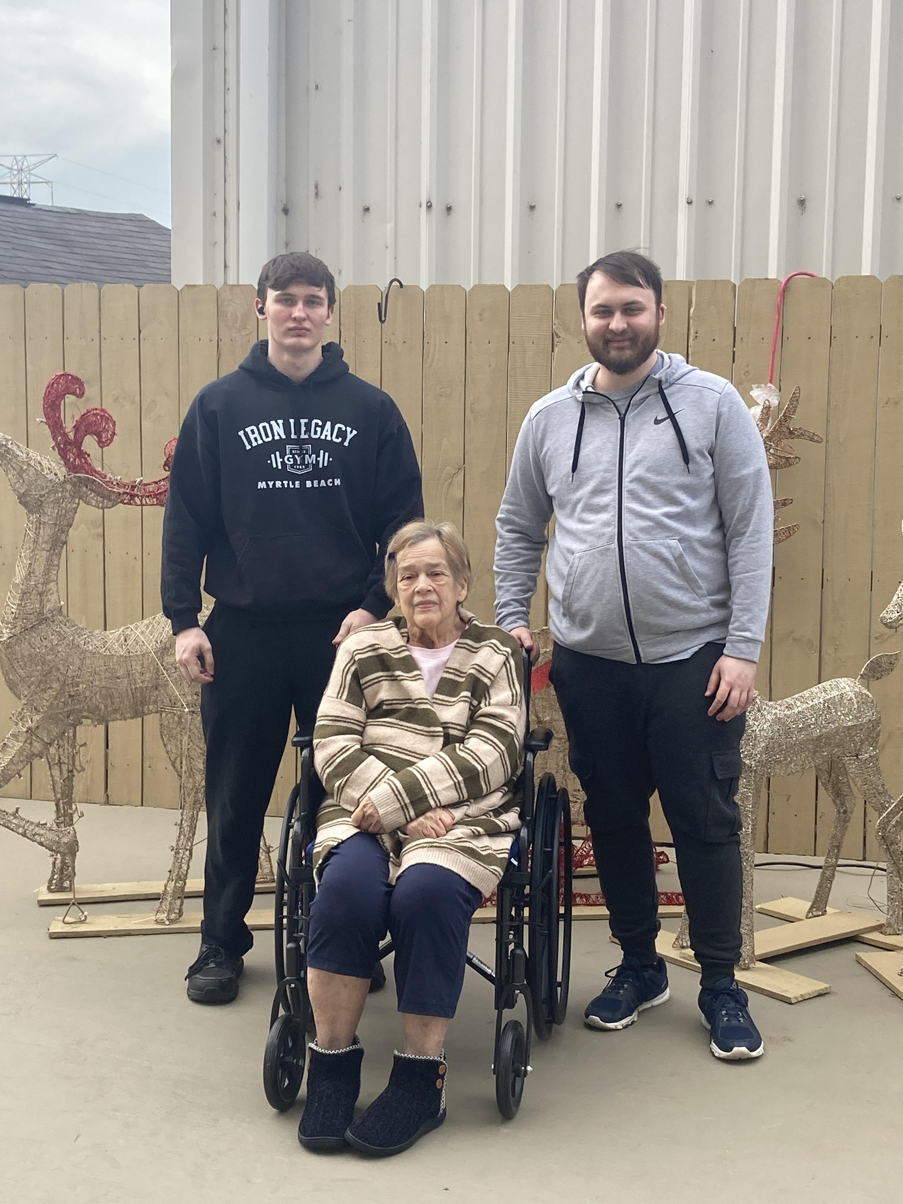 Two young men stand beside a woman in a wheelchair, smiling in front of holiday decorations.