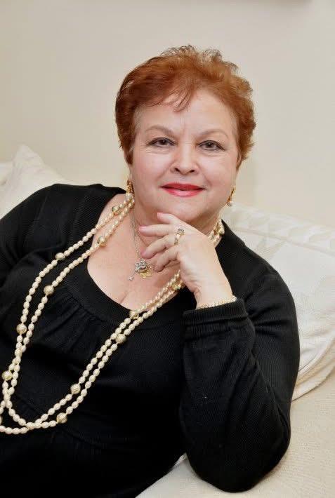 Woman with red hair sits elegantly on a couch, adorned with a pearl necklace, smiling warmly.