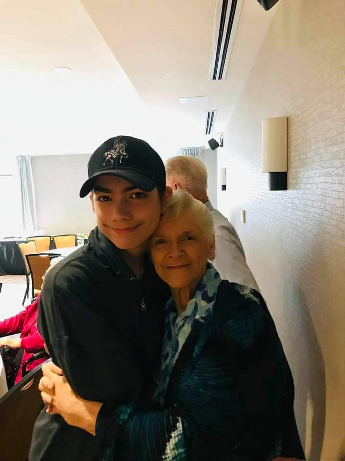 A young man warmly hugs an elderly woman in a cozy, softly lit indoor space.