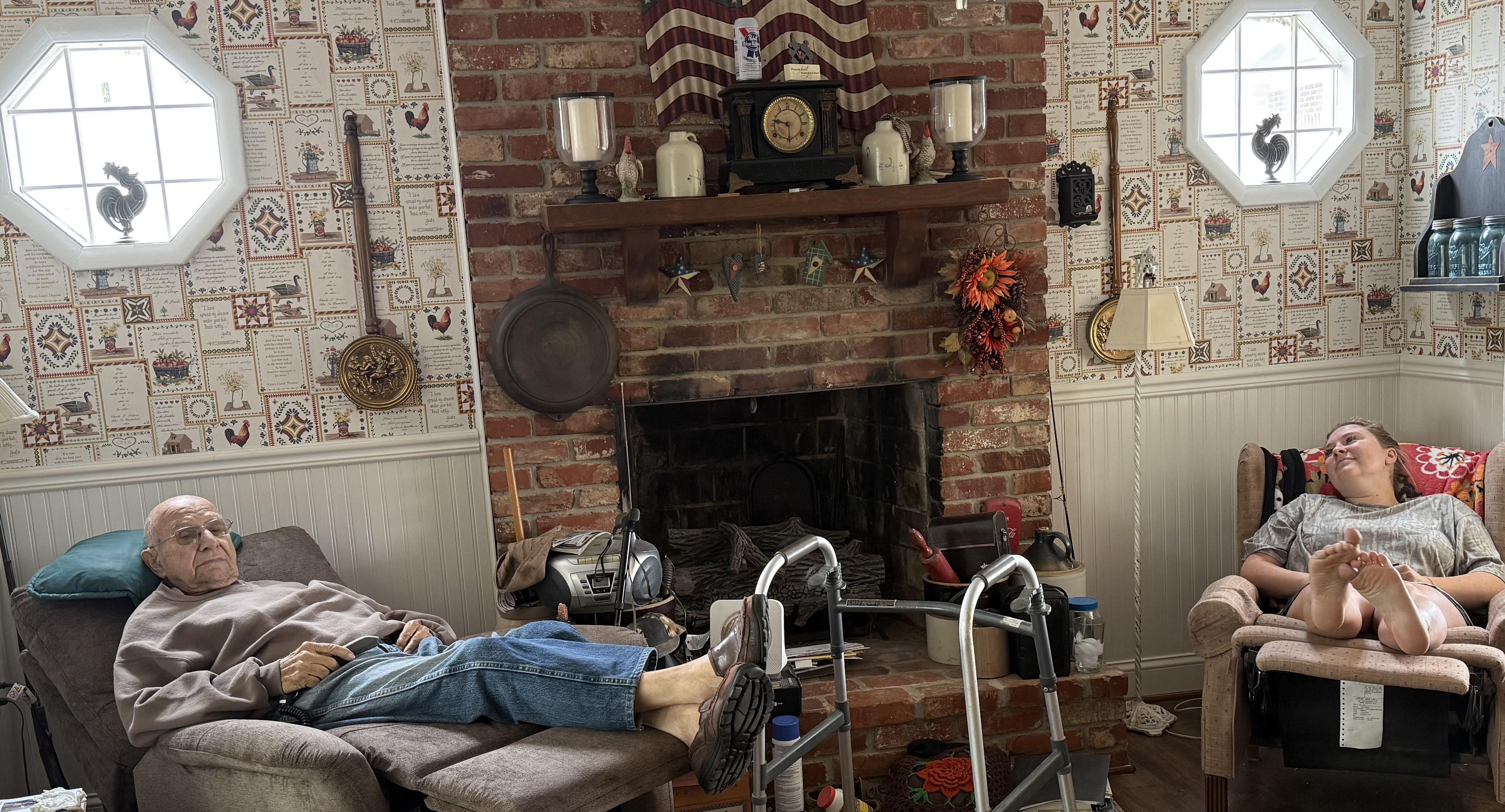 Two seniors relax in a cozy living room with a brick fireplace and warm decor.