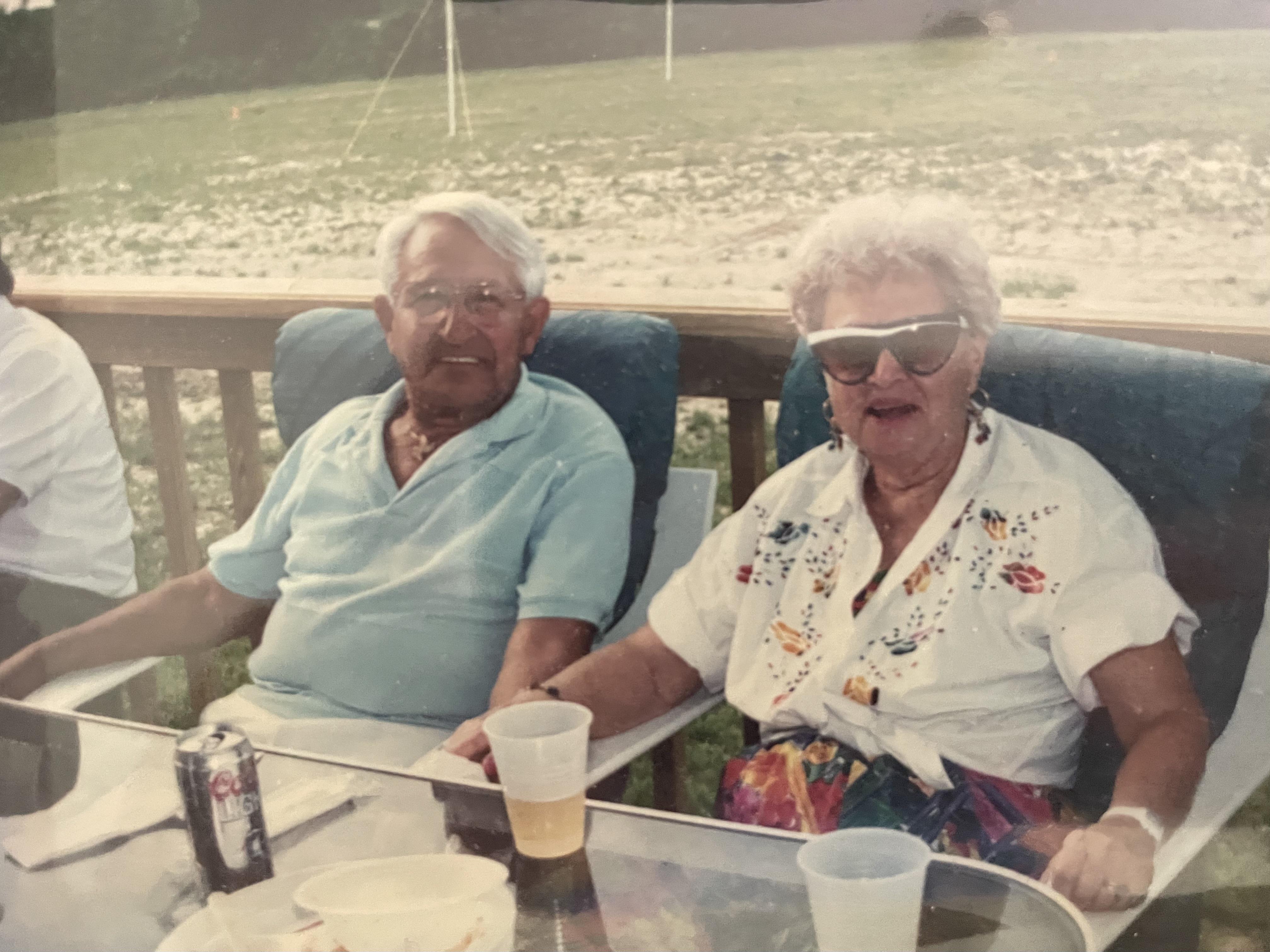 Two seniors sit at a table, enjoying drinks and each other's company outdoors.