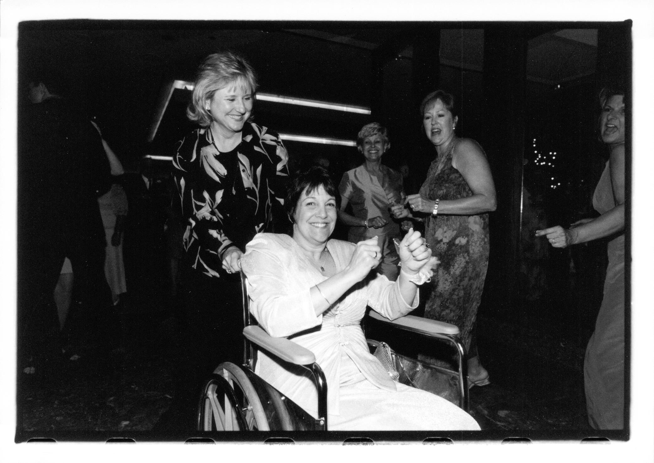 A group of friends dances and enjoys a lively celebration, with one woman in a wheelchair smiling.