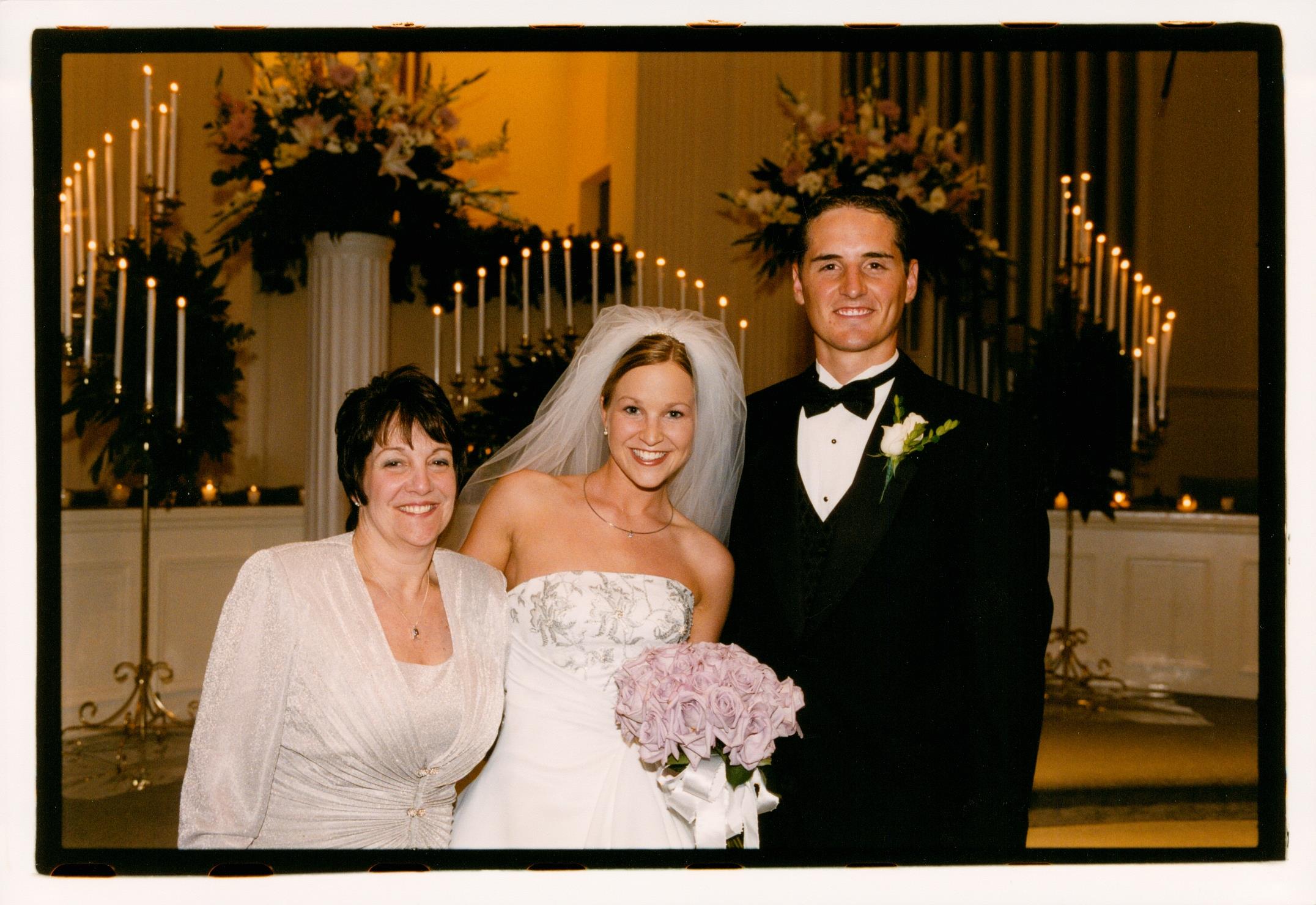 Couple in formal attire smiles with a family member at a wedding ceremony filled with decorations.
