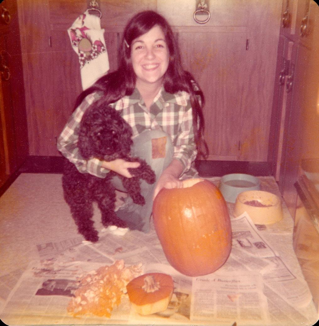 A woman carves a bright orange pumpkin with her black dog by her in a cozy kitchen.