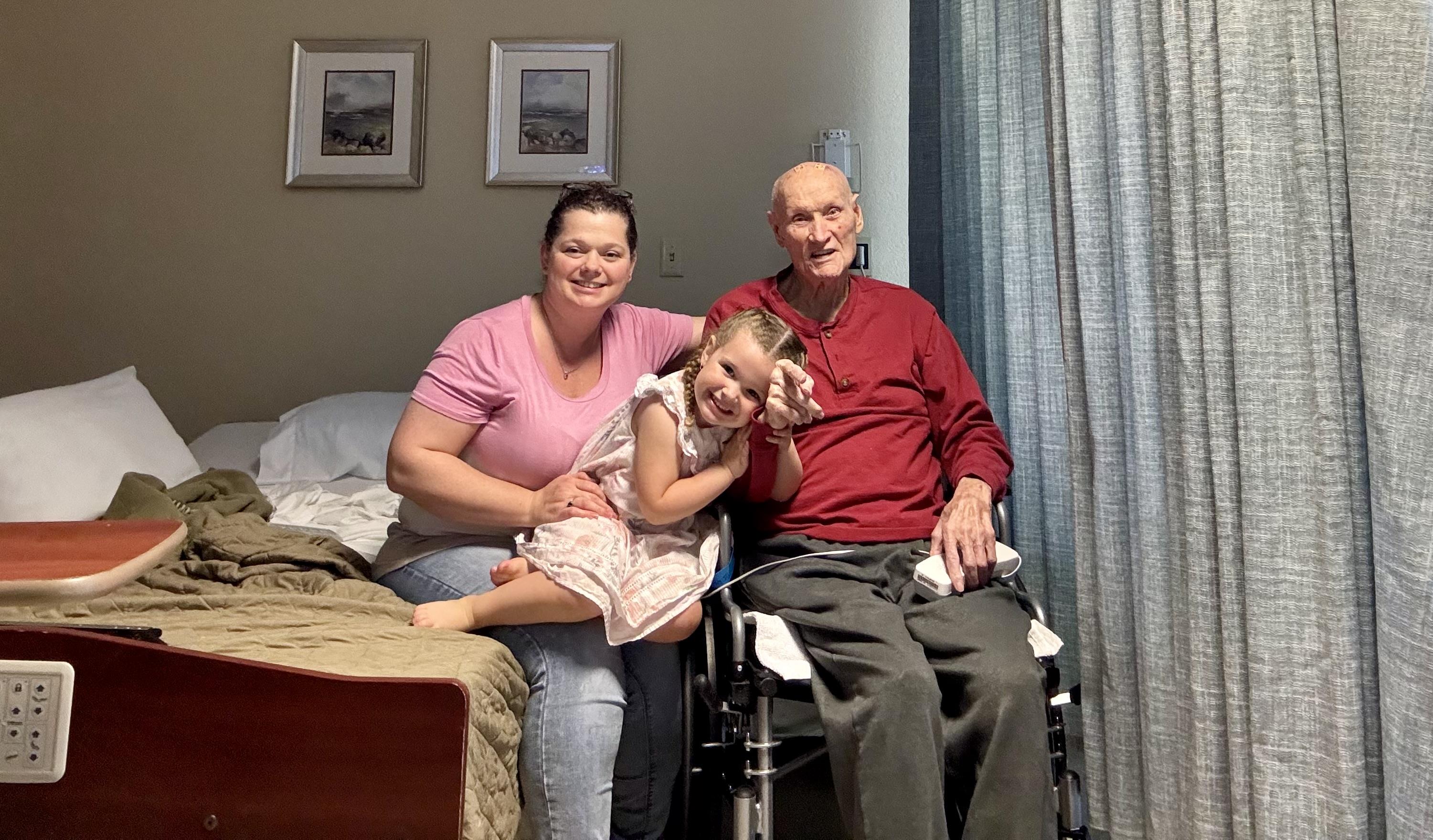 A mother and daughter smile warmly while sitting next to an elderly man in a hospital room.