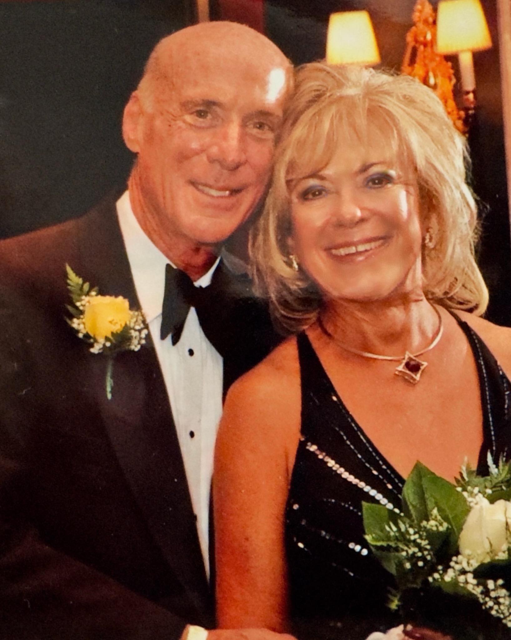 A mature couple smiles joyfully, showcasing their formal attire and enjoying a festive atmosphere.