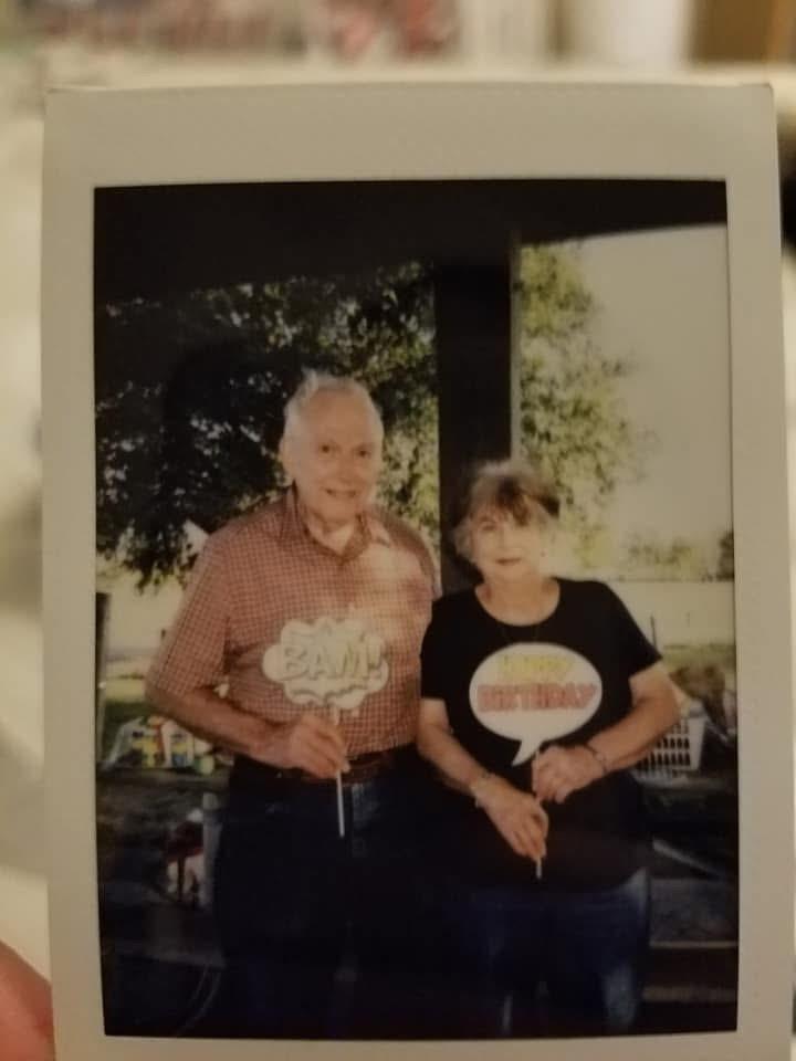 Elderly couple holds playful signs, enjoying a cheerful moment outdoors in bright sunlight.