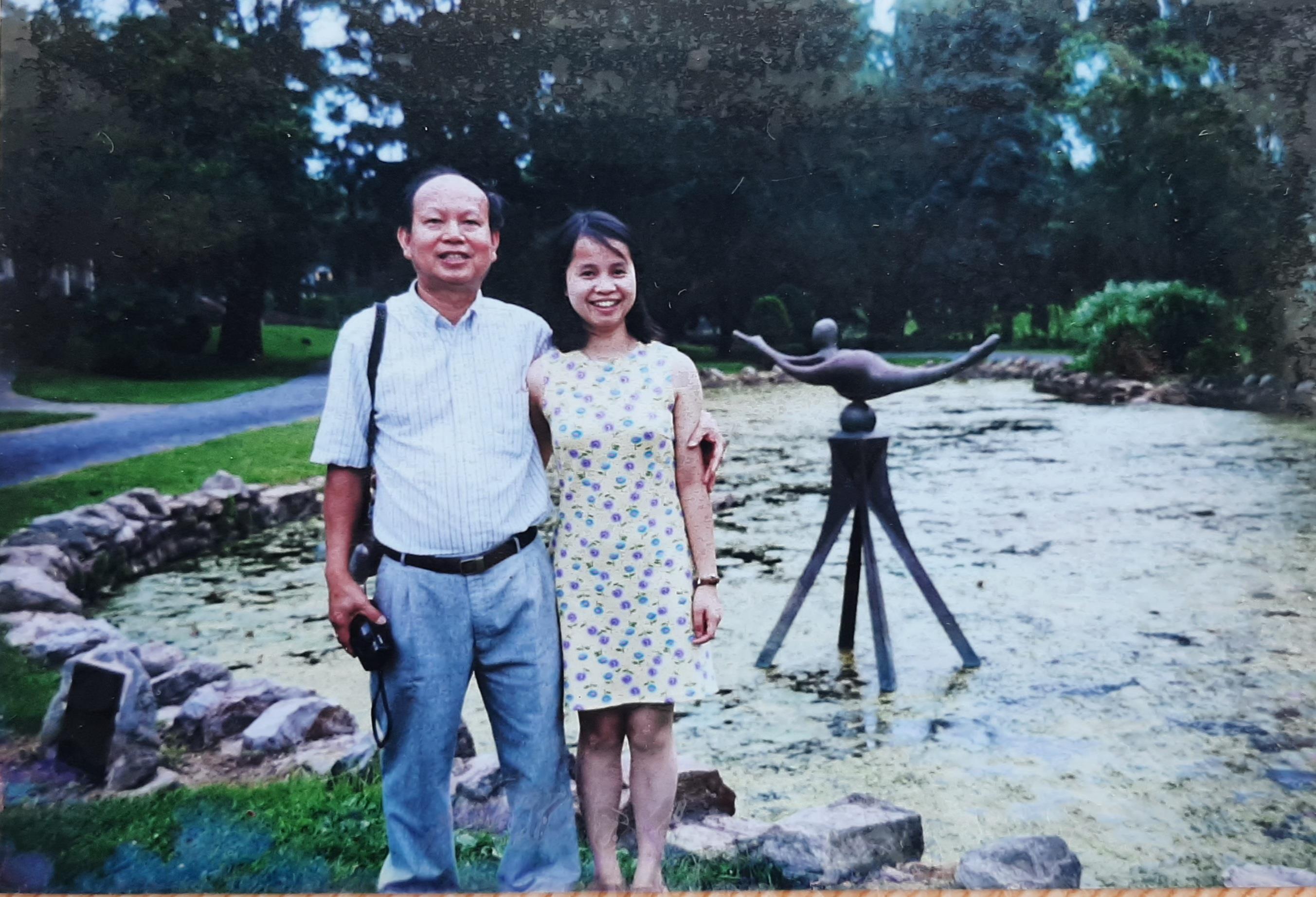 Two individuals pose happily by a pond adorned with a statue in a park.