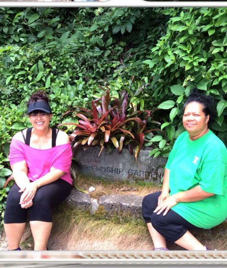 Two friends sit together on a stone bench surrounded by vibrant greenery and plants.