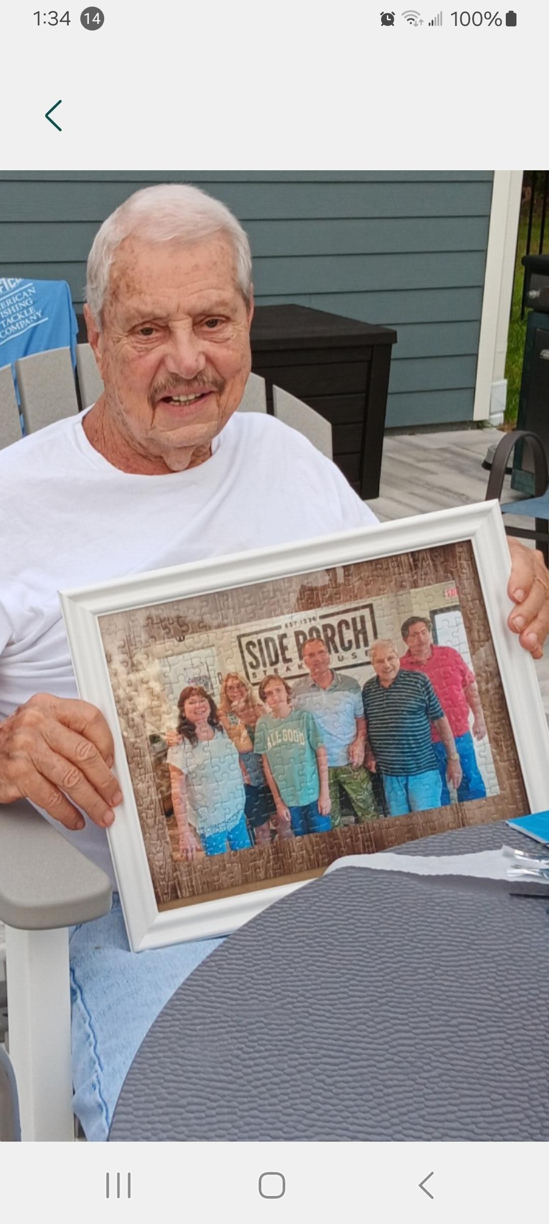 An elderly man smiles while holding a frame with a family photo during a summer gathering.