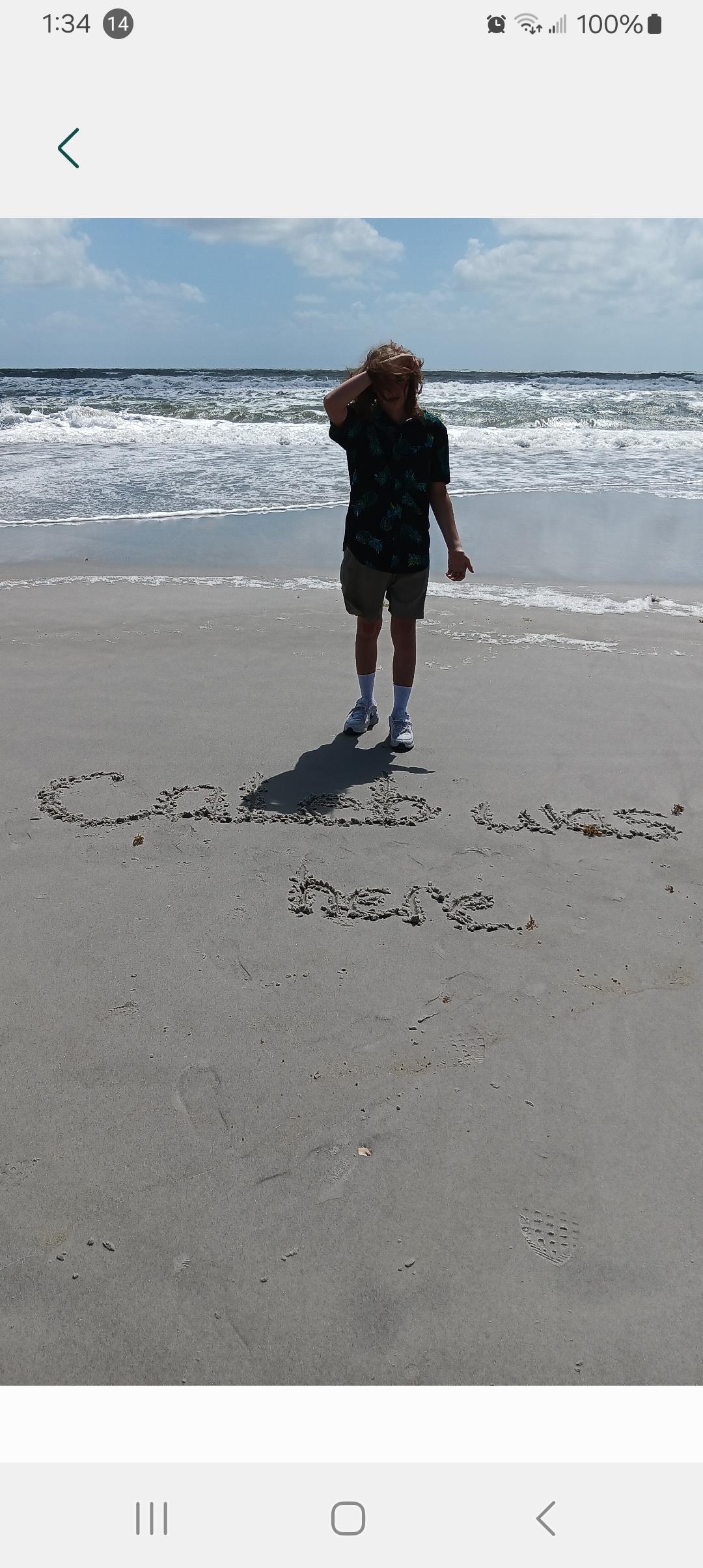 Individual stands on sandy beach, brushing hair back, beside words inscribed in sand.