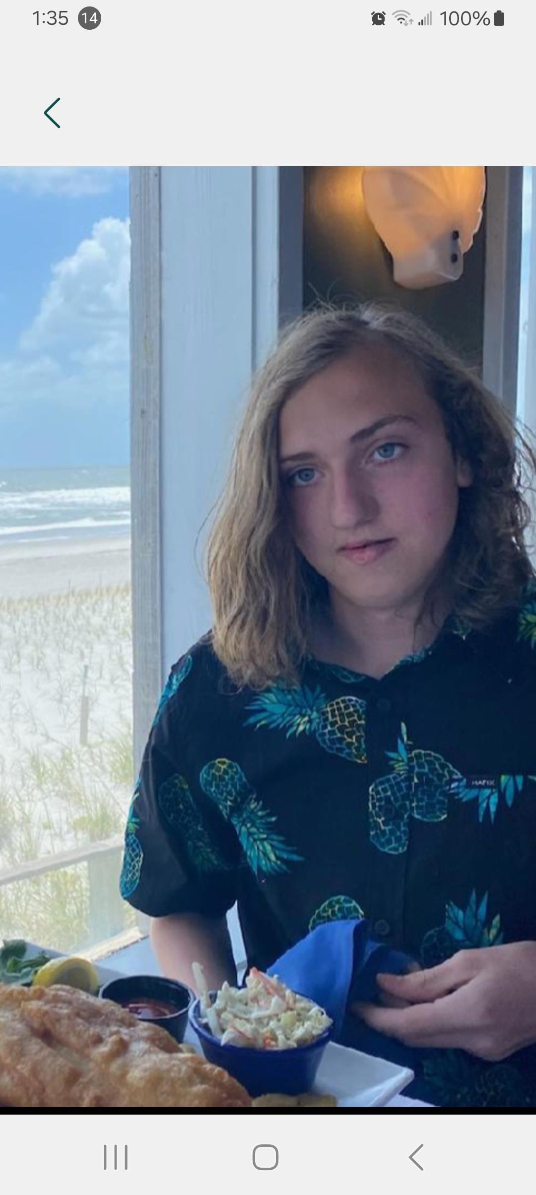 A person stands indoors, holding food and looking out at the beach with clouds in the background.