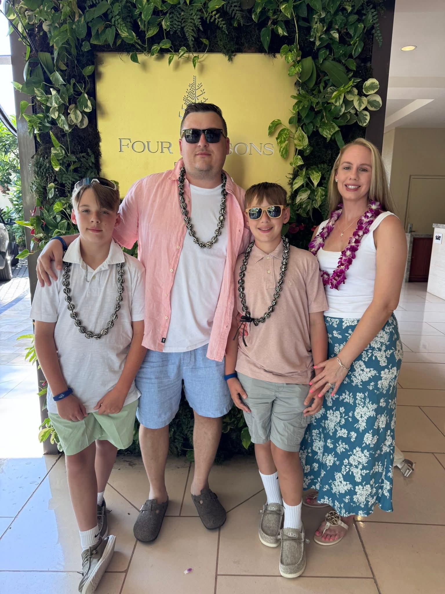 Family poses together at a resort entrance, dressed in casual vacation clothing and leis.