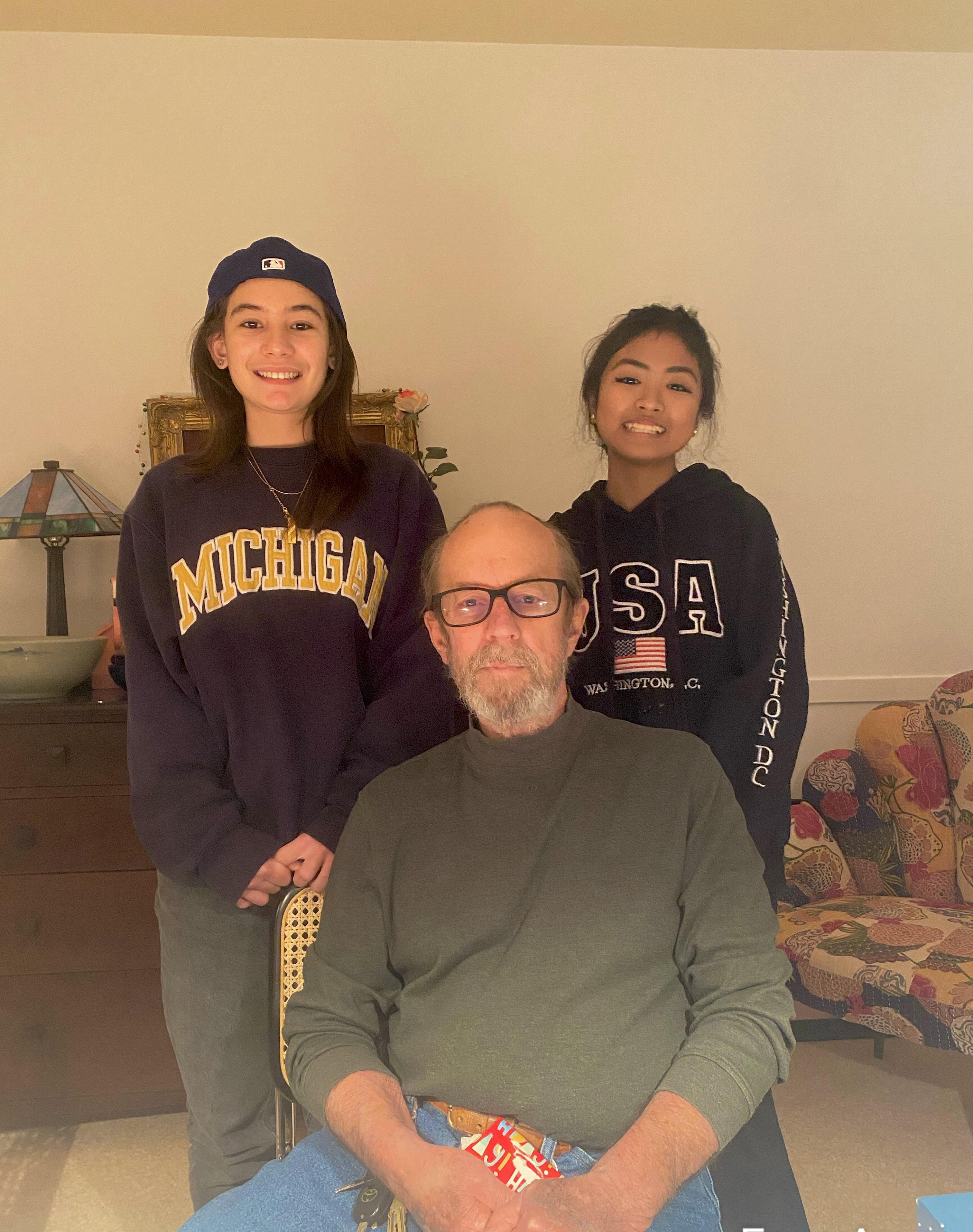 Two young women stand beside a man seated in a comfortable living room, all smiling.