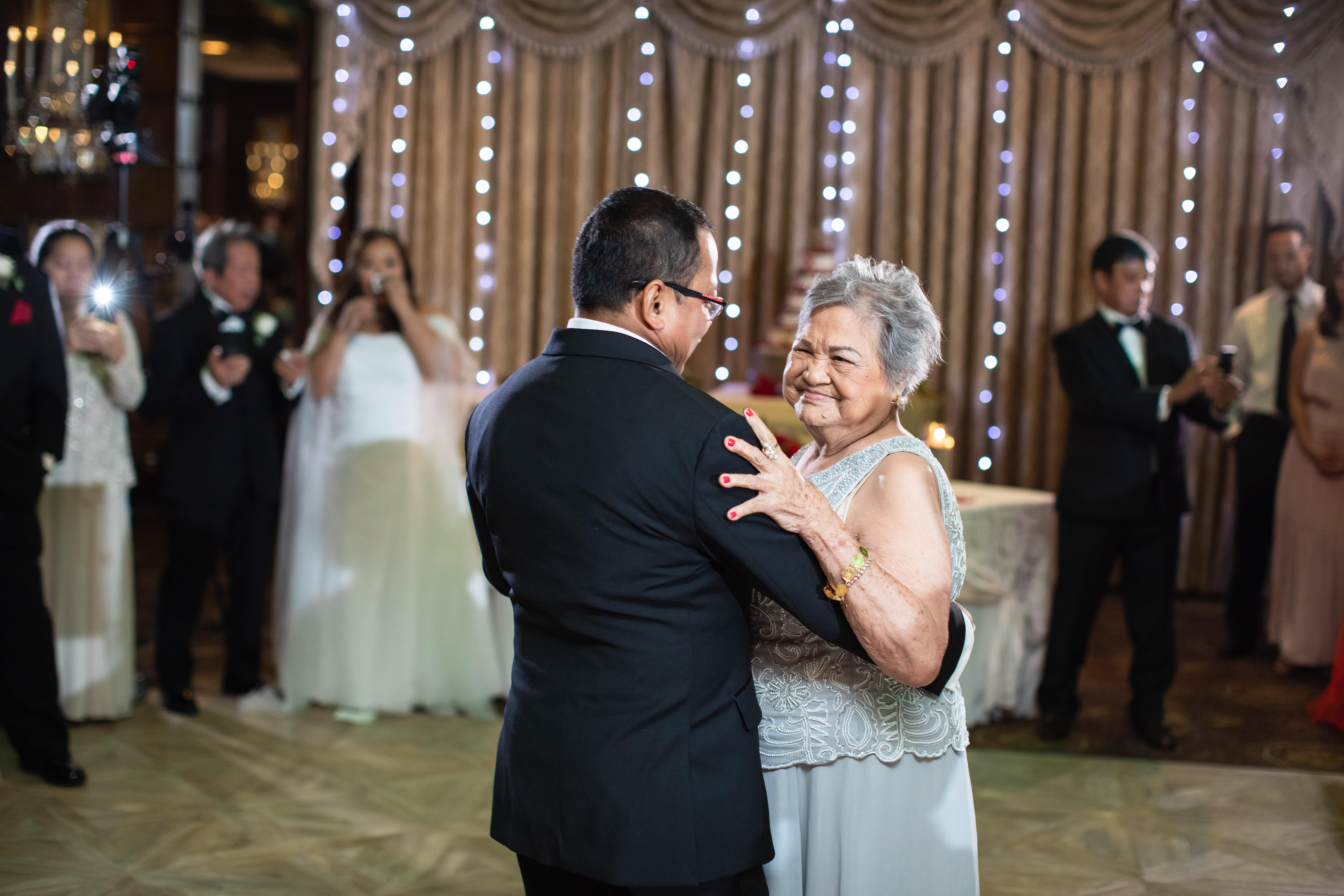 A man and an elderly woman dance together, surrounded by family and festive lights.