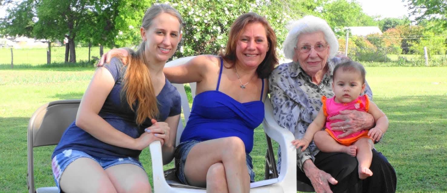 Four women of different ages sit together in a backyard surrounded by greenery, sharing smiles.