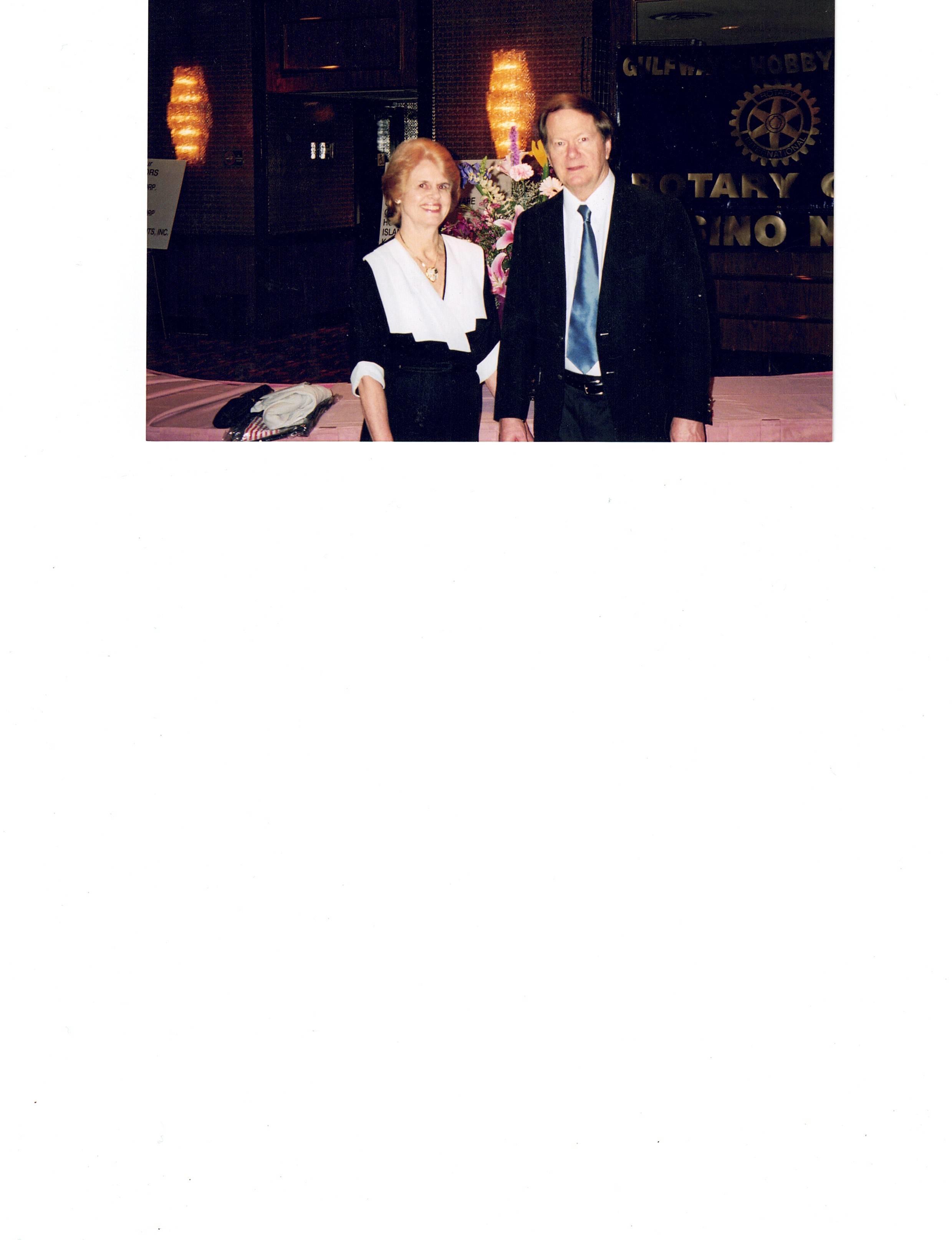 A couple stands together, dressed elegantly for a celebration at a festive venue in the evening.