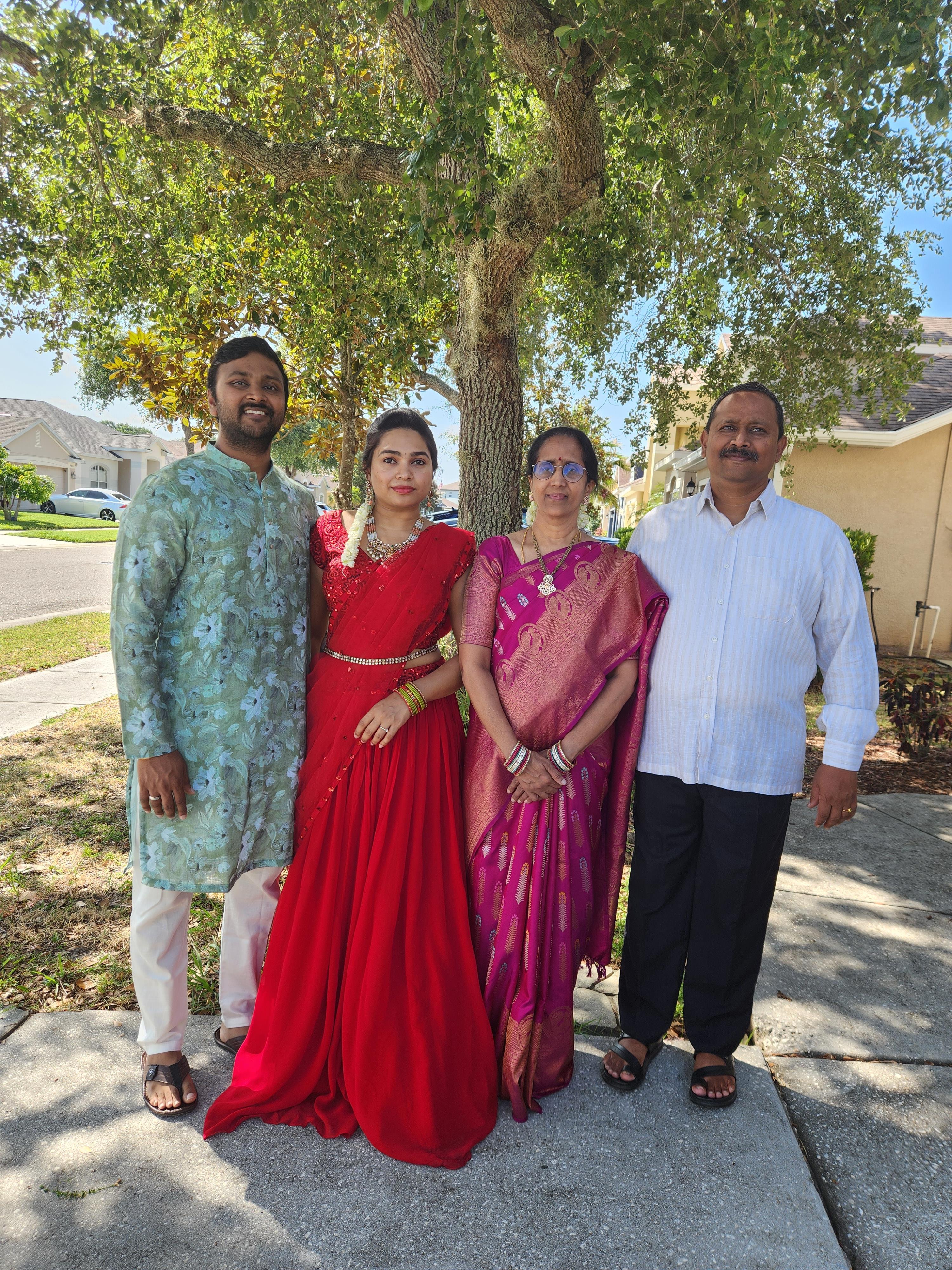 Four people in colorful traditional attire stand outside a house on a sunny day.