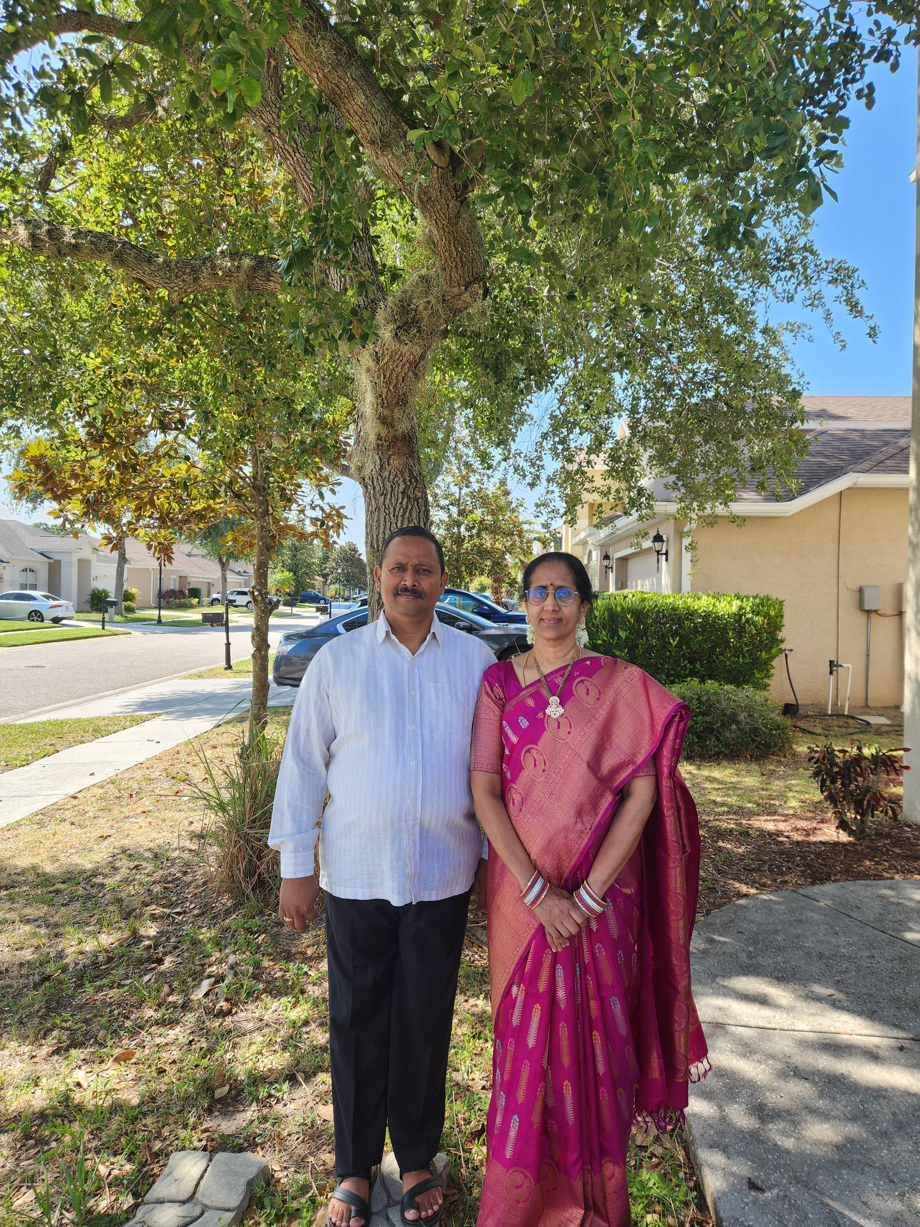 A man and woman wearing traditional clothing stand happily by a tree in a quiet residential area.
