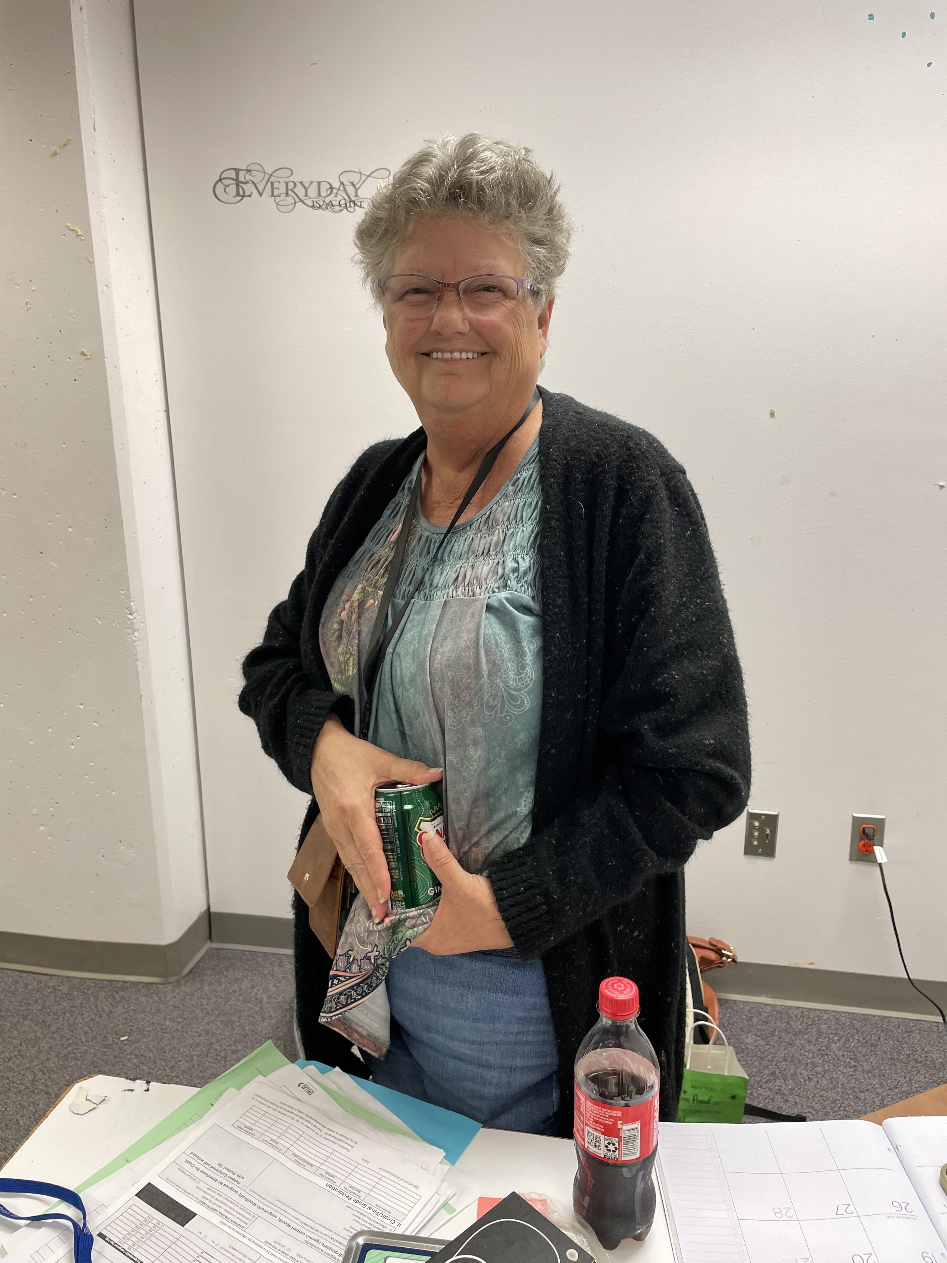 A cheerful woman stands in a casual setting with a drink and paperwork, engaging warmly with others.