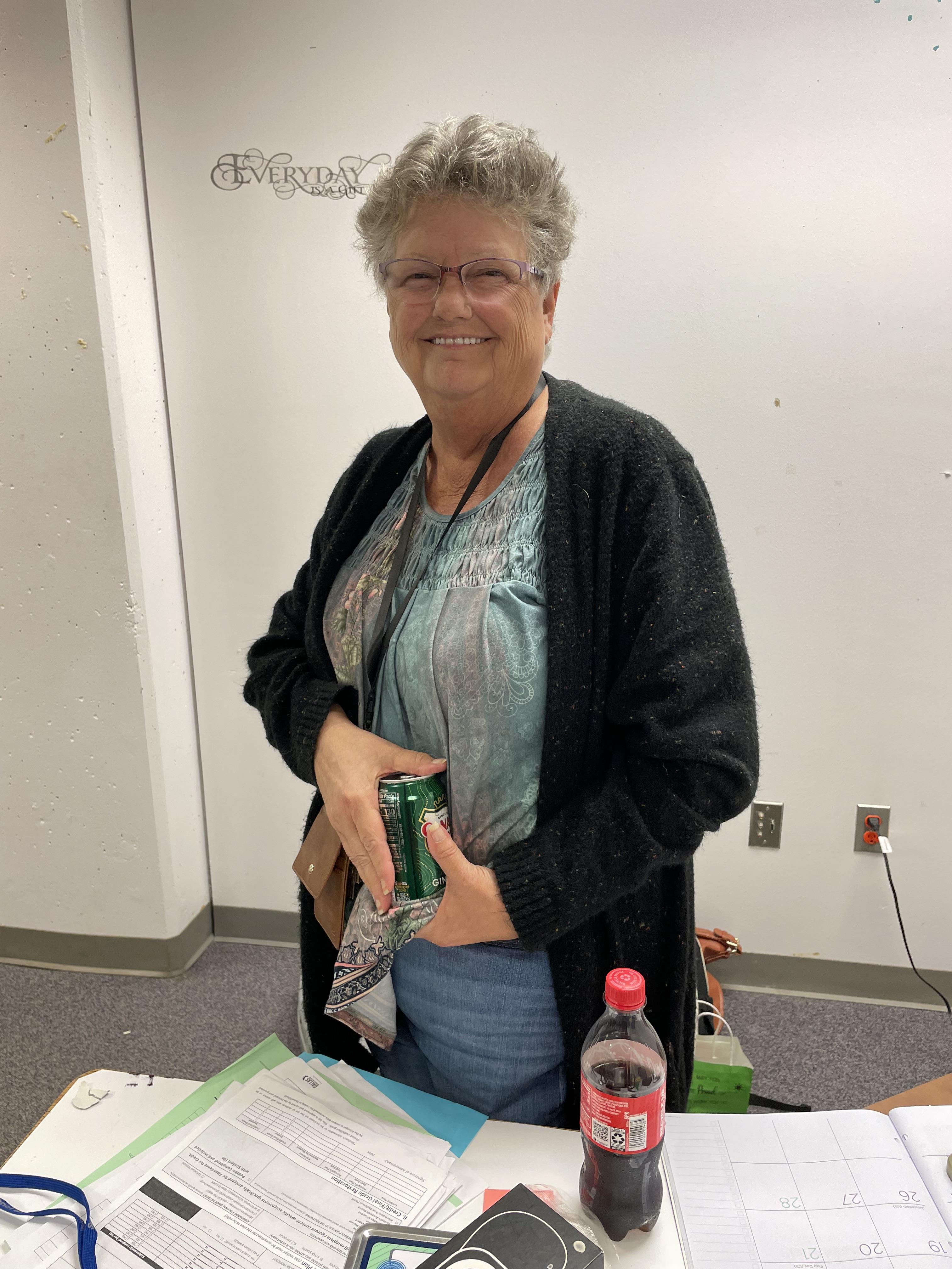 A woman with gray hair smiles warmly while holding a soda can in a community center setting.