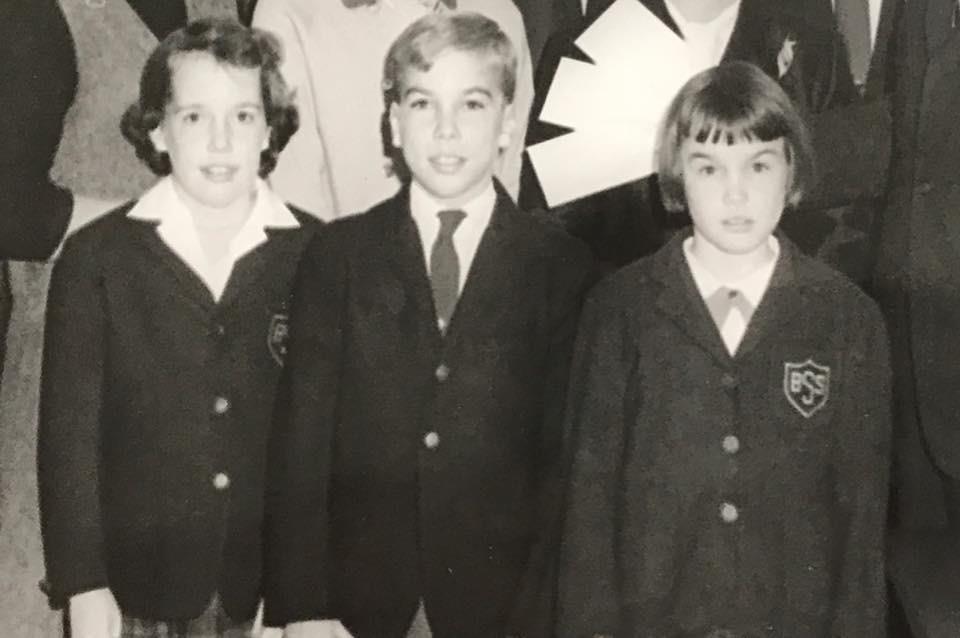 Three children in black school uniforms stand together, smiling for a group photo in a classroom.