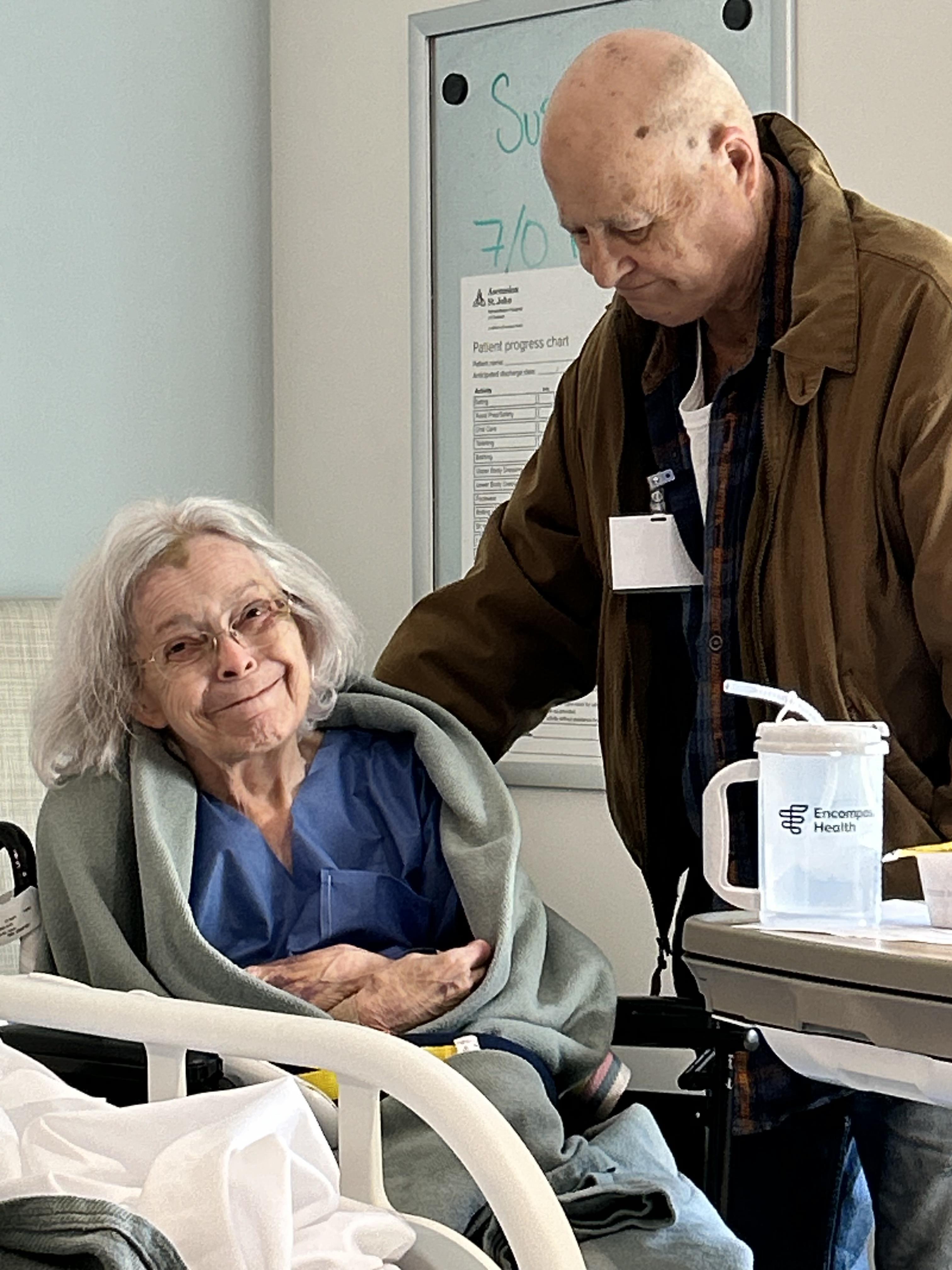 An elderly man comforts a smiling woman wrapped in a blanket while sitting in a hospital.