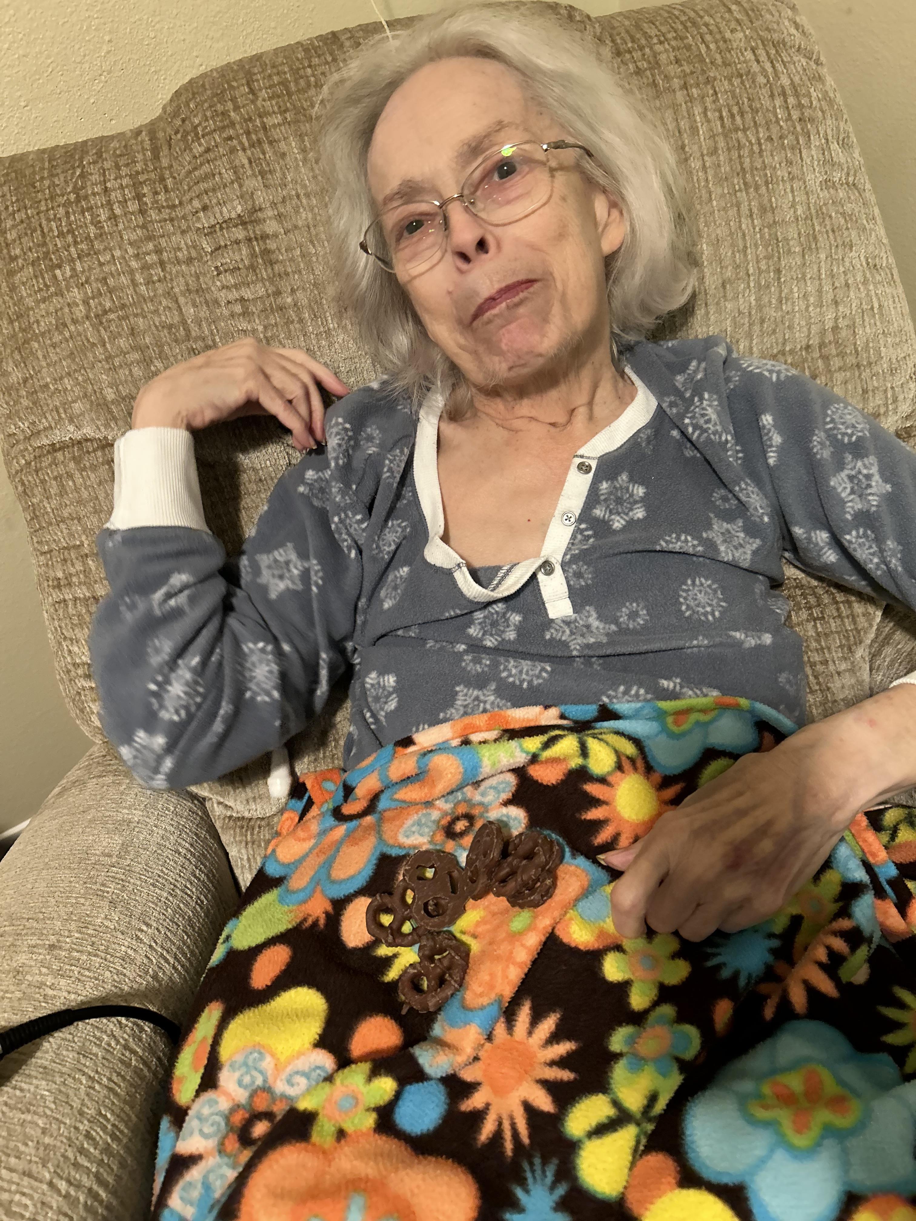 An elderly woman smiles gently while cozy in a chair, wrapped in a colorful blanket.