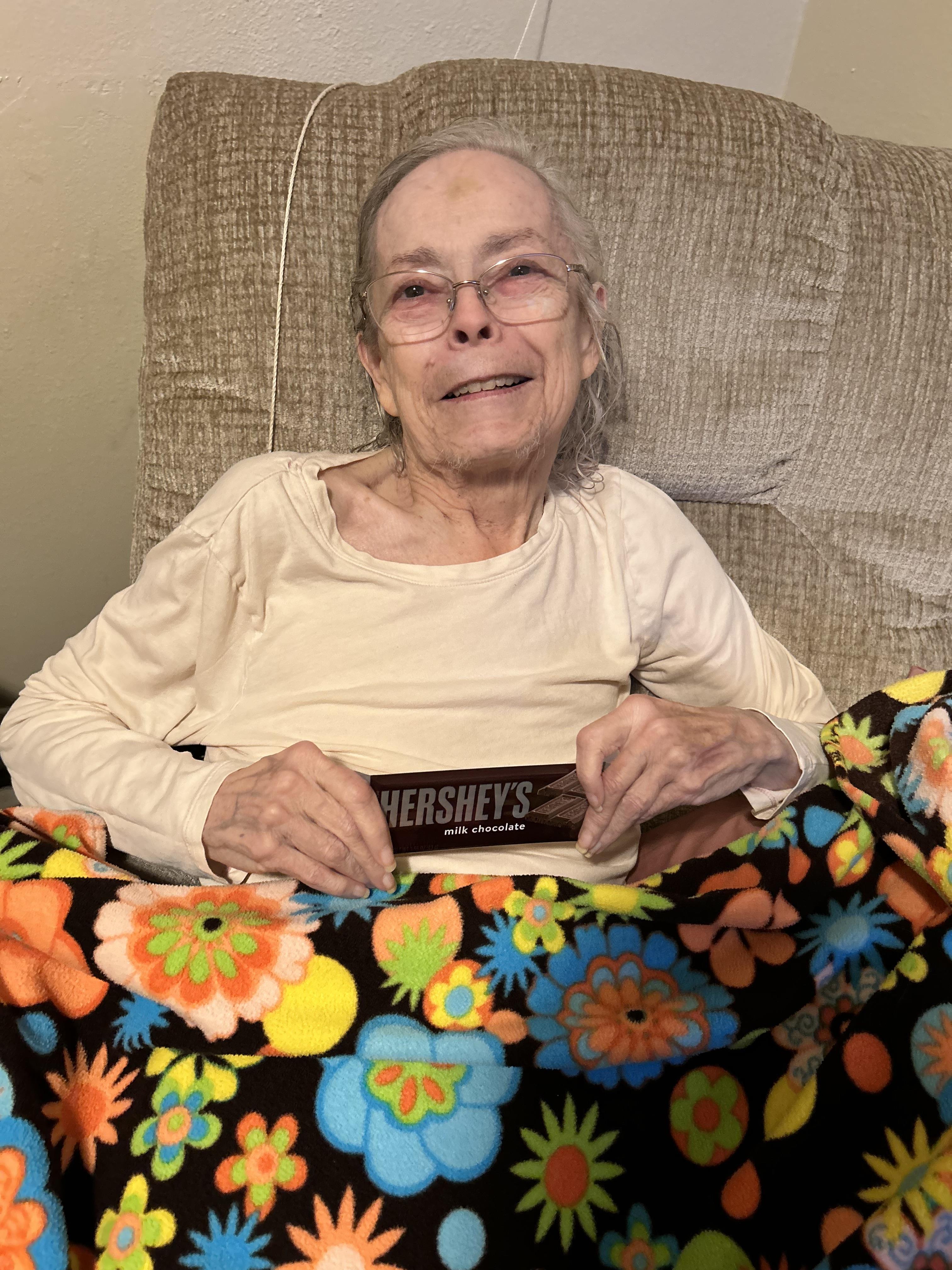 An elderly woman enjoys her time sitting in a cozy chair while holding a chocolate bar.
