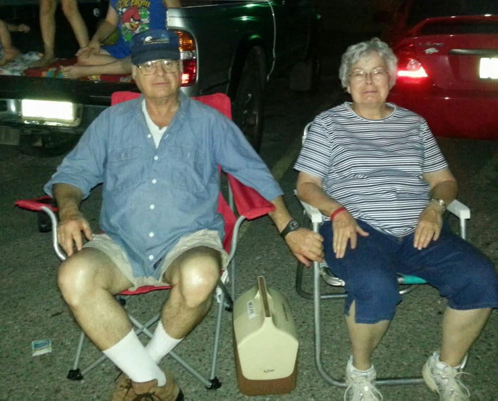 Older couple sits in camping chairs at a neighborhood gathering, holding hands and chatting.