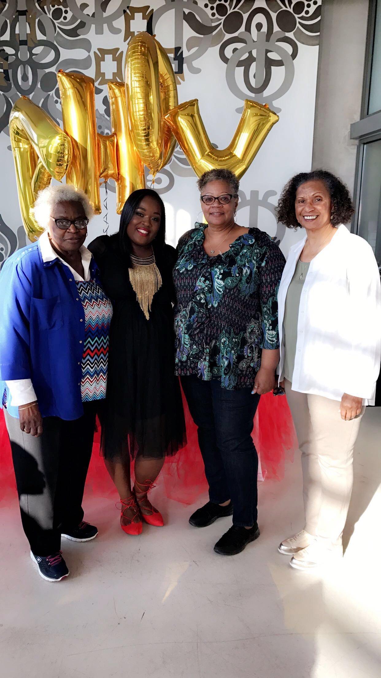 Four women gather indoors to celebrate together, posing cheerfully with large gold balloons.
