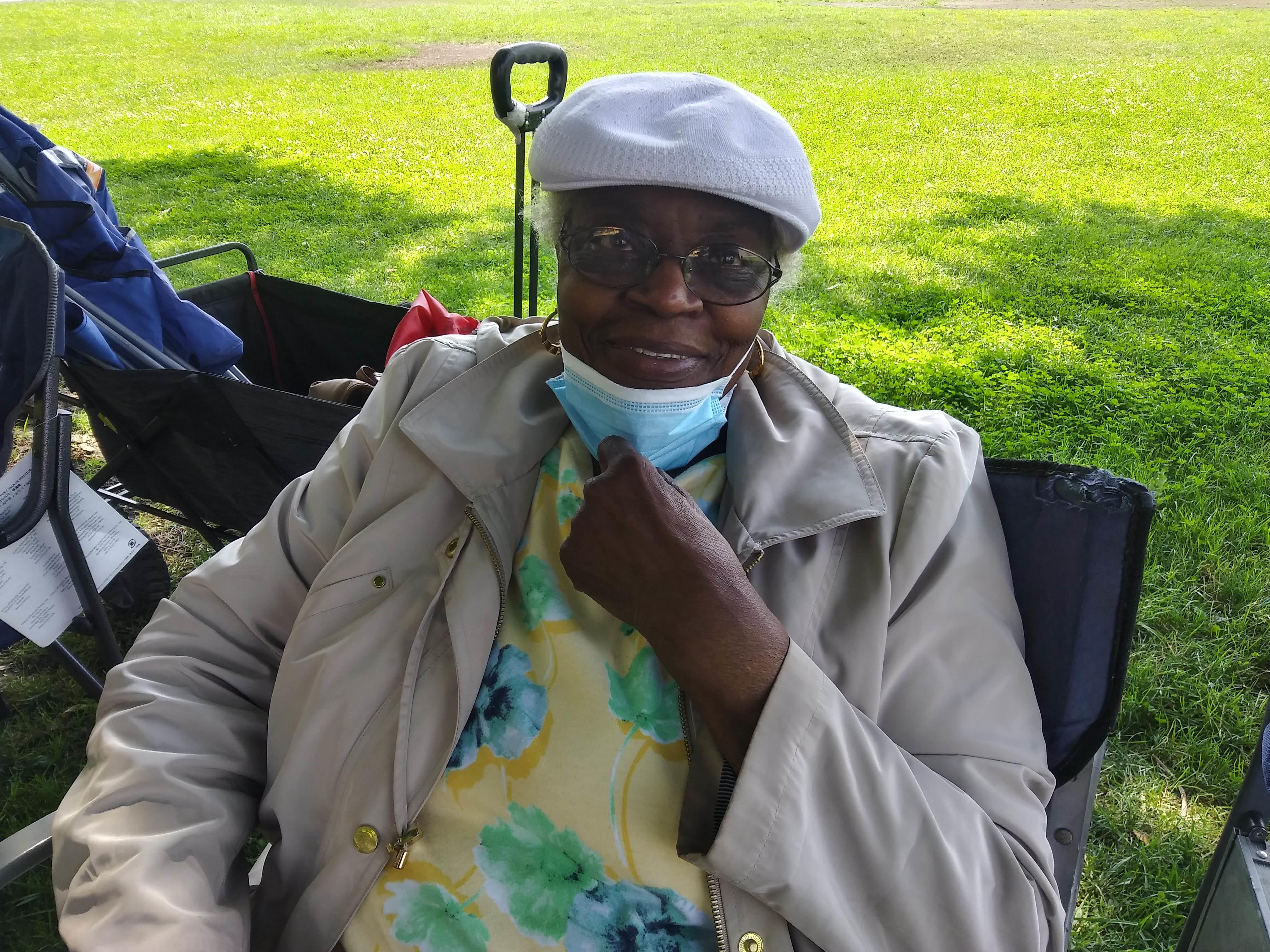 Woman with glasses and a hat sits on a chair in a park enjoying a sunny afternoon with greenery.
