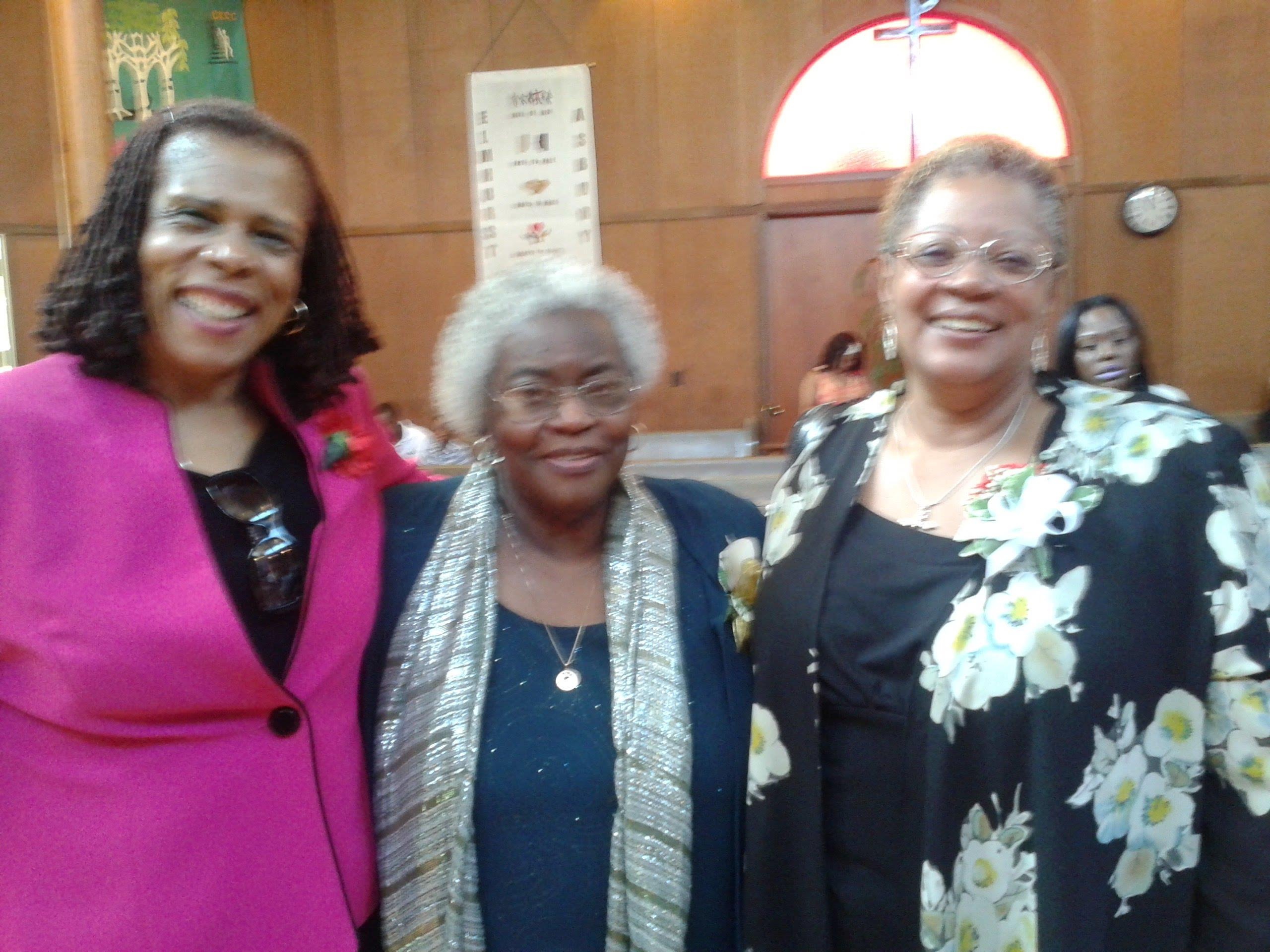 Three women happily pose at a community event, flaunting their vibrant outfits.