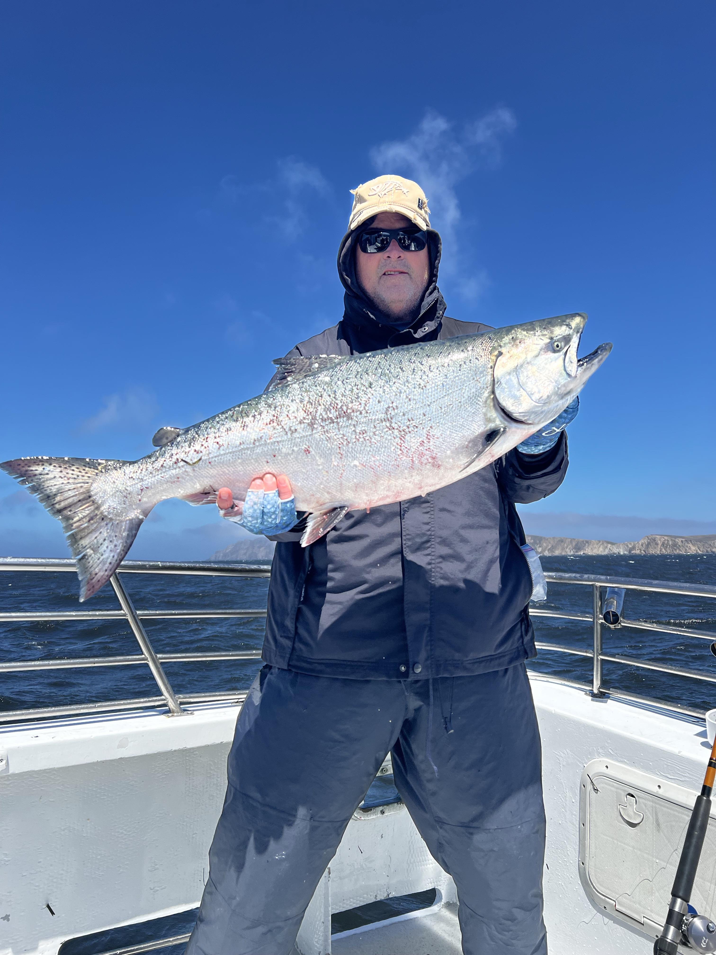 Fisherman holds a big fish while standing on a boat under clear blue skies.