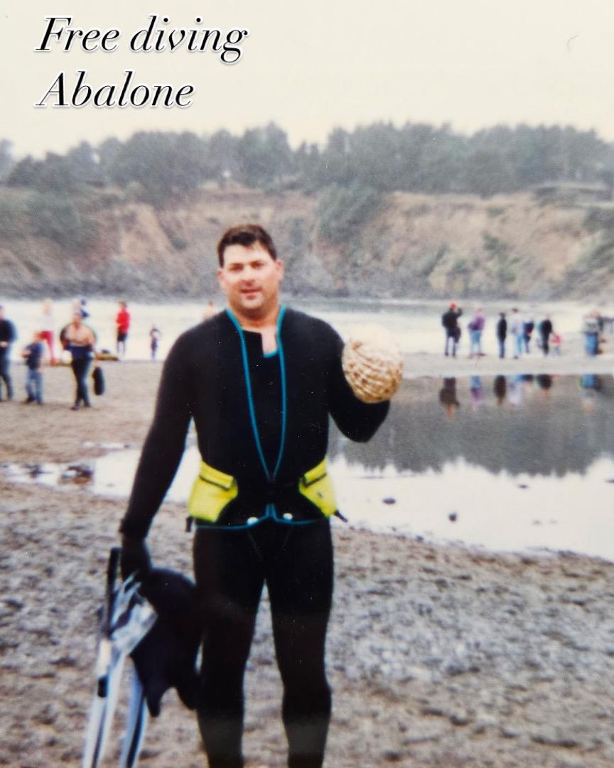 Diver wearing a wetsuit proudly displays abalone at a coastal diving site with spectators nearby.