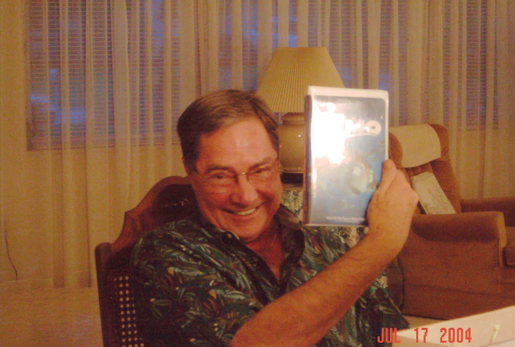 A man with glasses shows a VHS tape while smiling in an inviting living room.