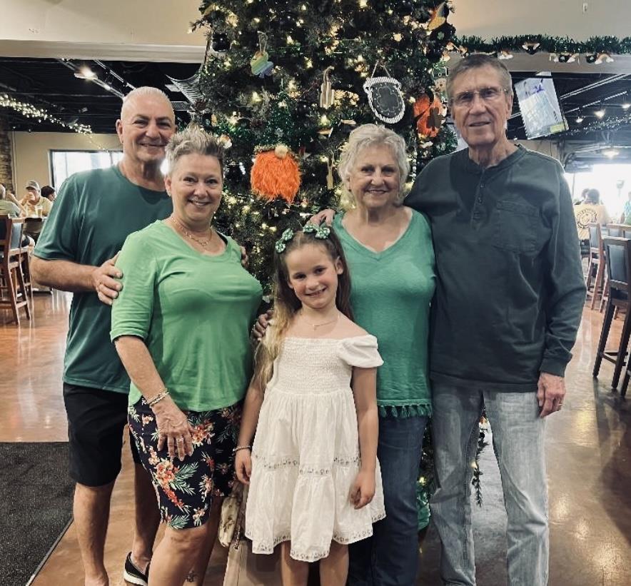 Five family members pose together next to a brightly decorated tree in a lively restaurant.