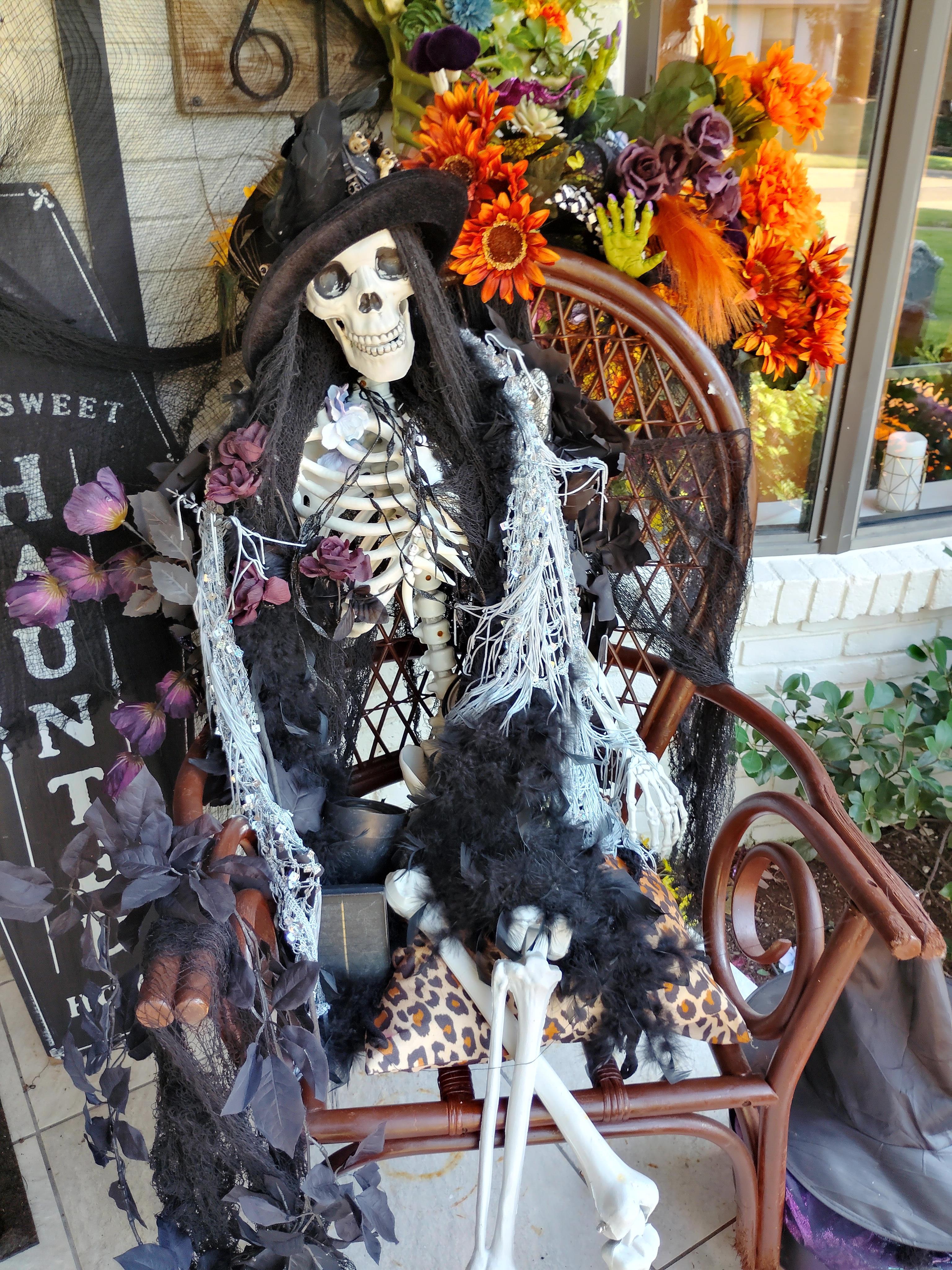A skeleton dressed as a witch sits on a chair adorned with autumn decorations.