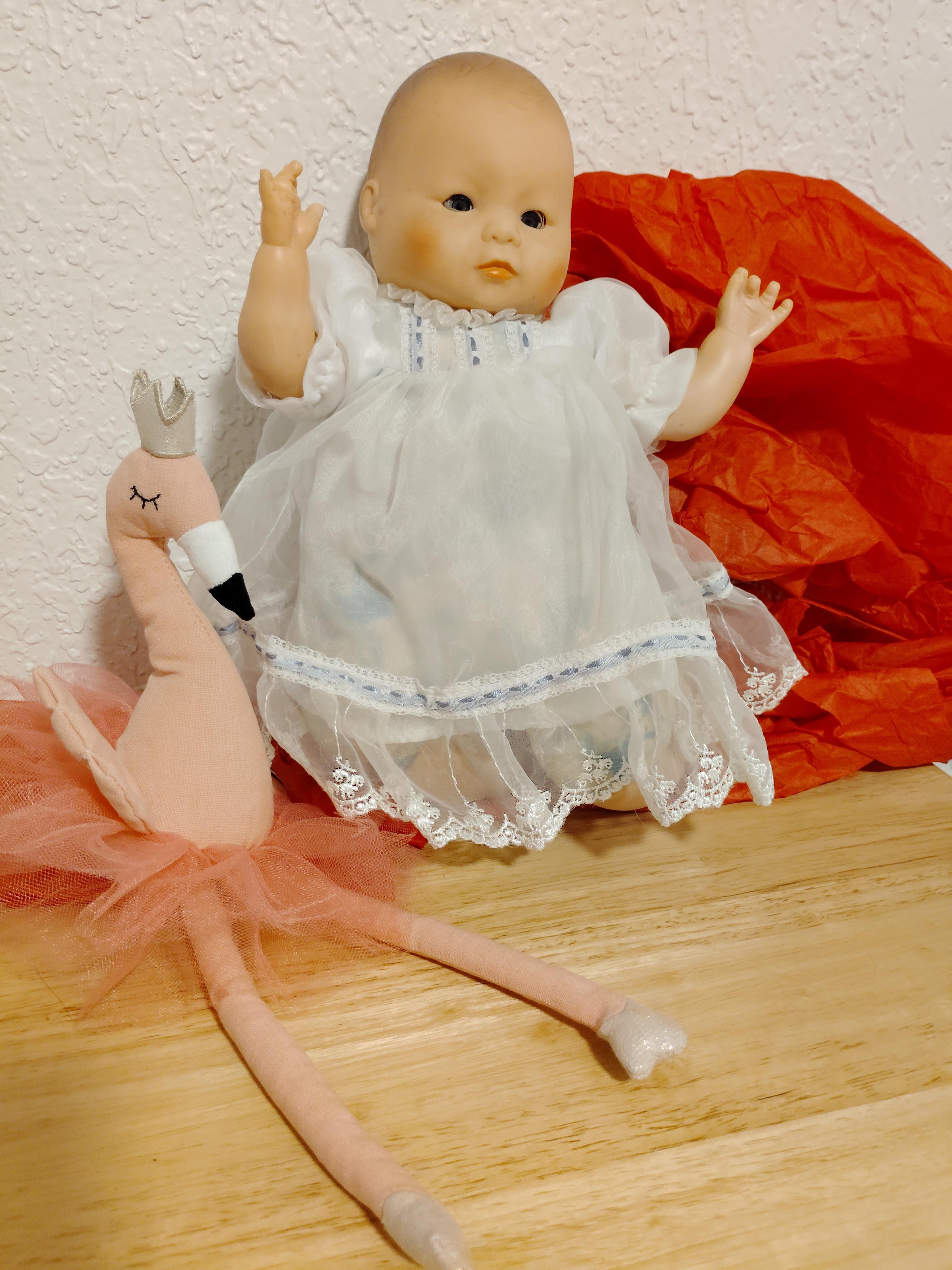 A doll in a white dress and a flamingo toy are on a wooden table with red paper behind them.