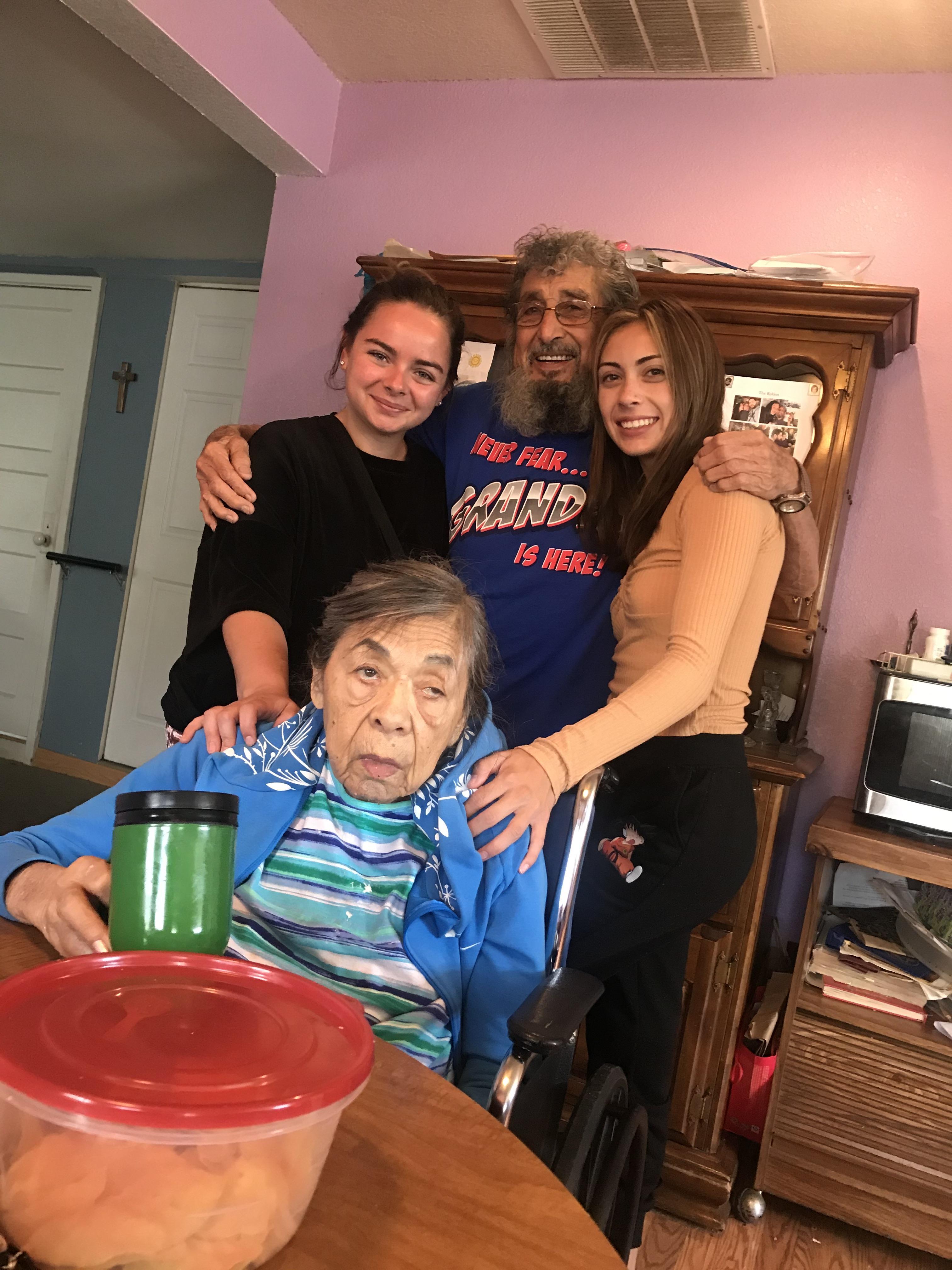 Three young adults embrace a smiling elderly man while seated at a table in a warm living space.