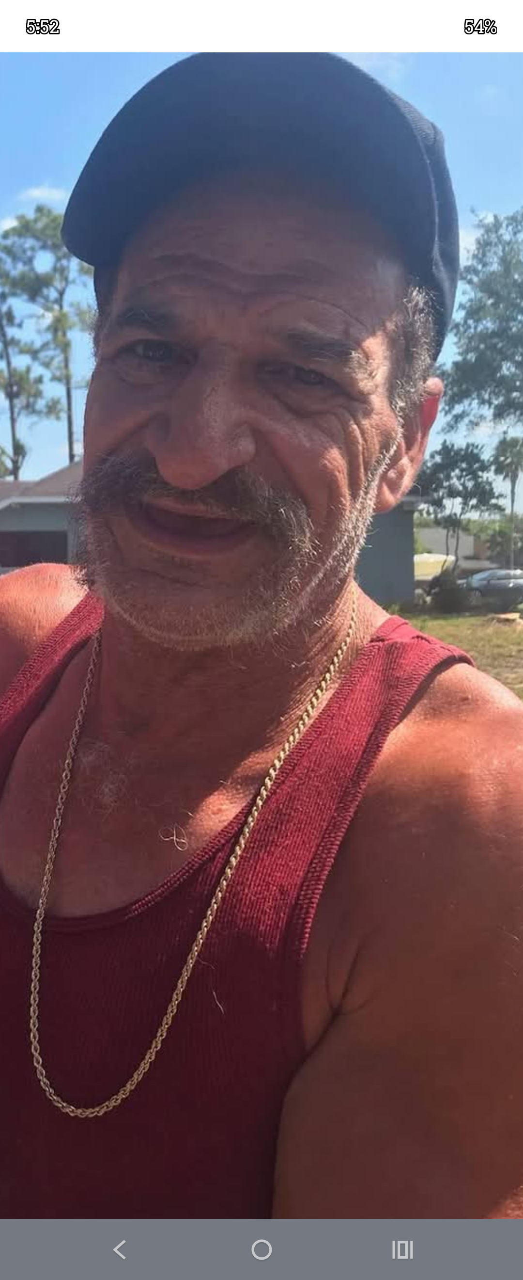 A man with a mustache smiles in a red tank top under bright sunlight. Trees surround him.