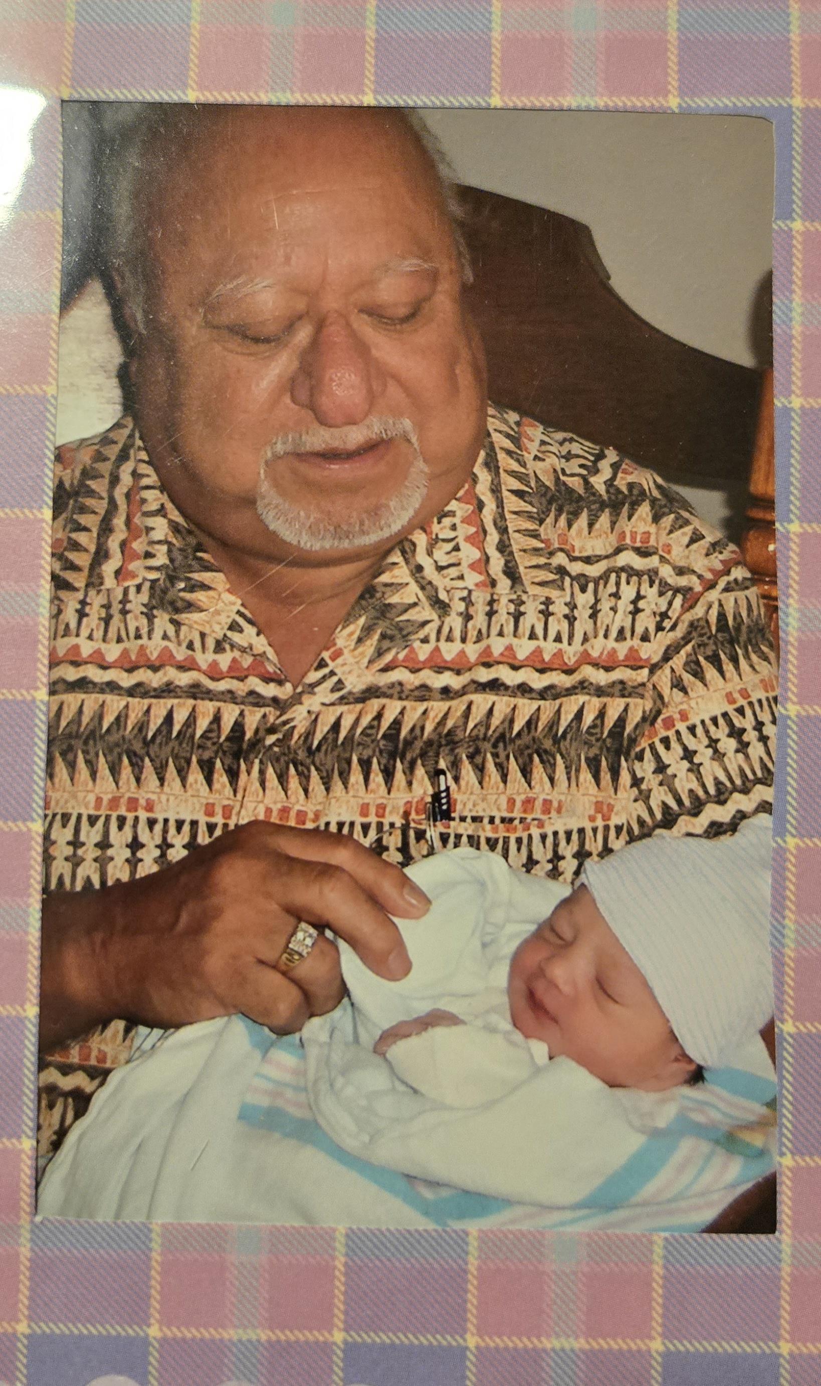 A grandfather smiles softly as he holds his sleeping grandchild in a cozy home setting.