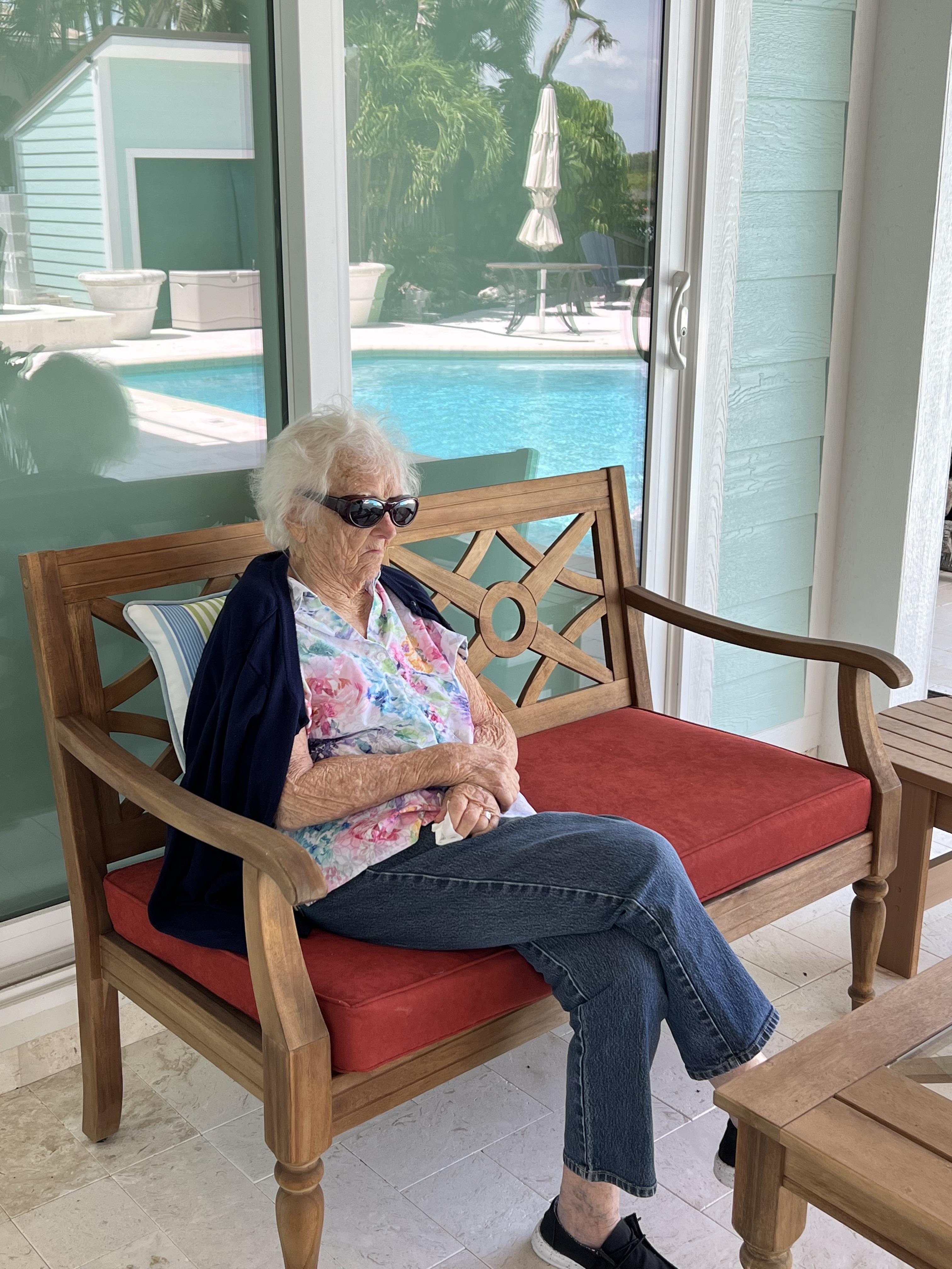 Senior woman enjoys a sunny day on a patio sofa near a swimming pool while wearing sunglasses.