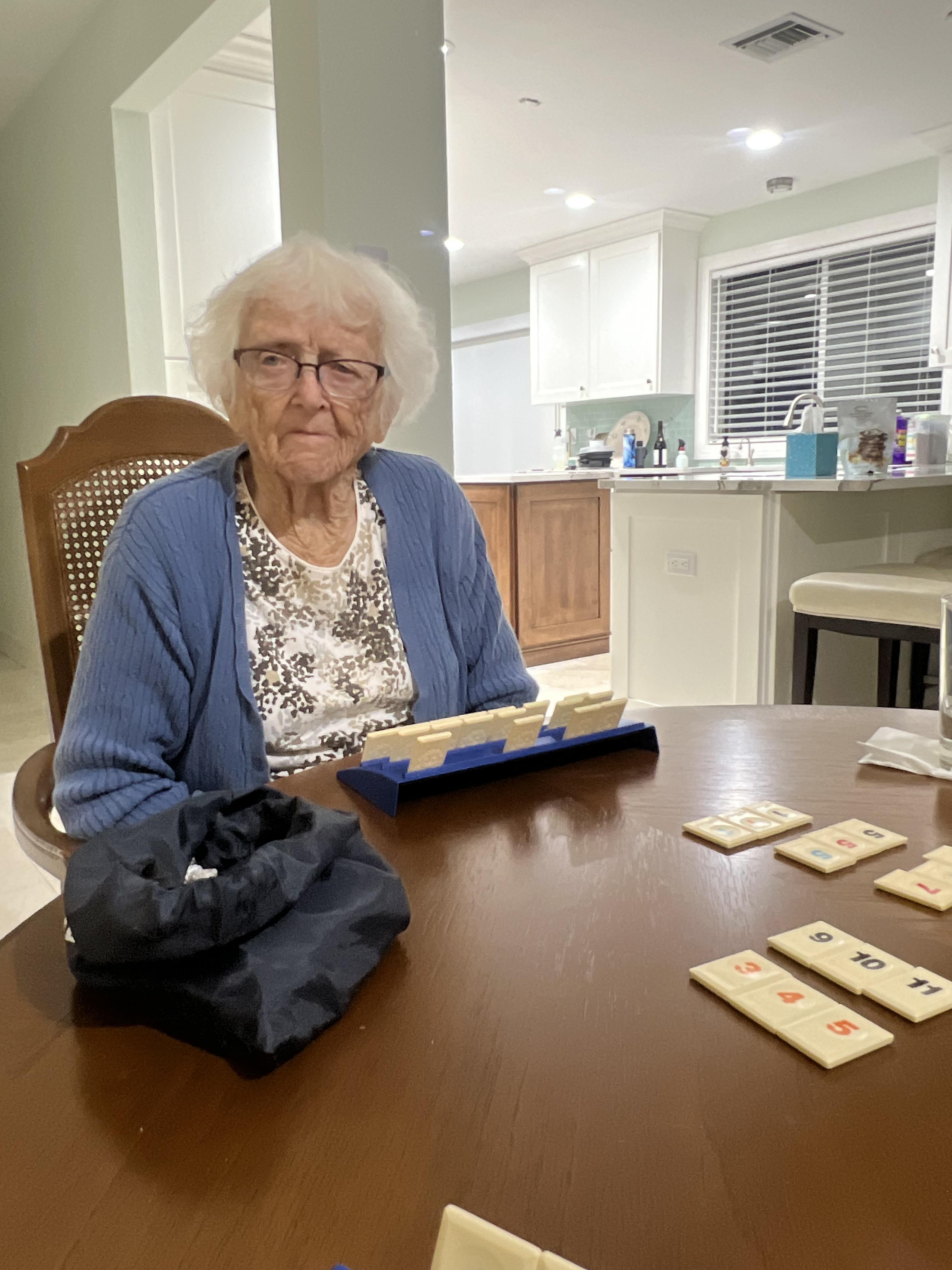An elderly woman sits at a wooden table, engaged in a board game at home.